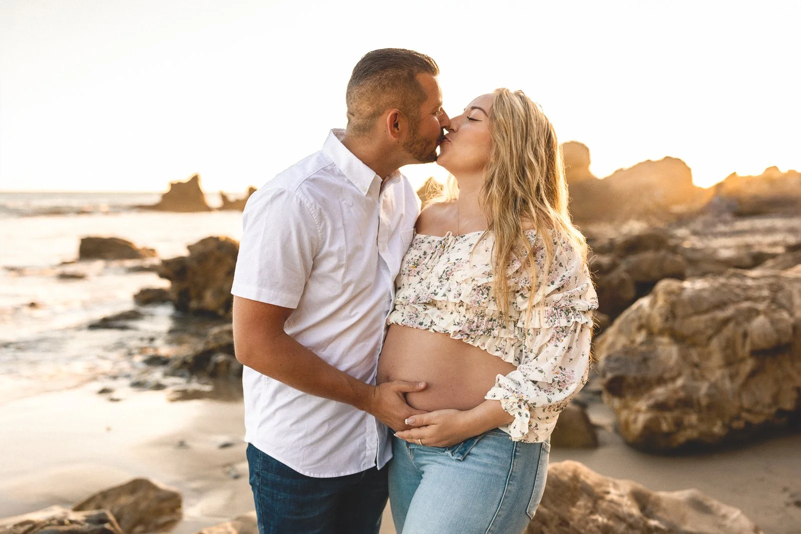 Couple kissing on a rocky beach during golden hour maternity photos.