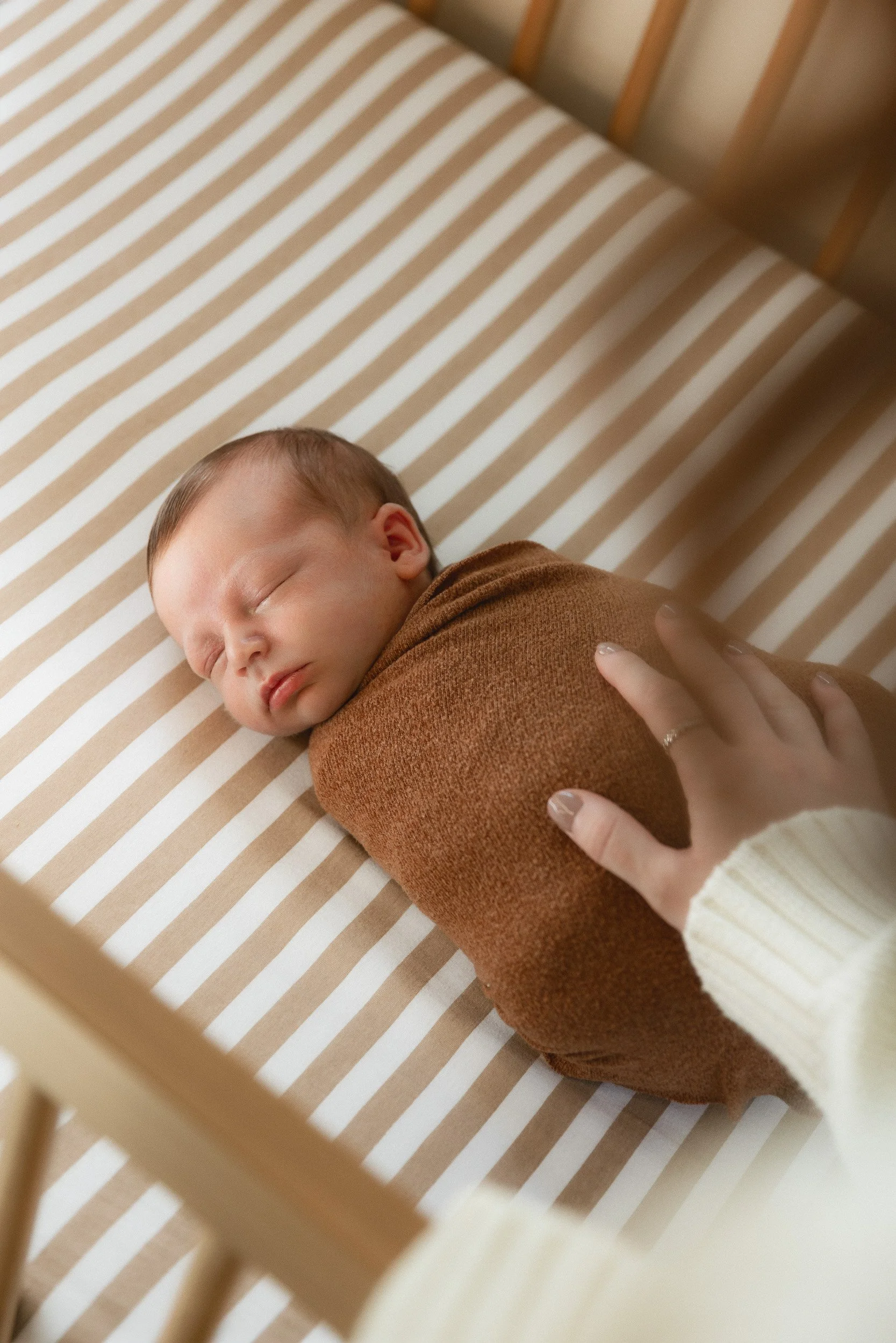 Swaddled newborn sleeping in a striped crib with a parent's hand resting on them during an in-home newborn session in Orange County