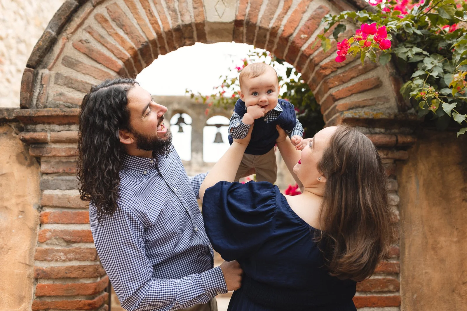Parents lifting their baby under a brick arch with flowers at Mission San Juan Capistrano in San Juan Capistrano.