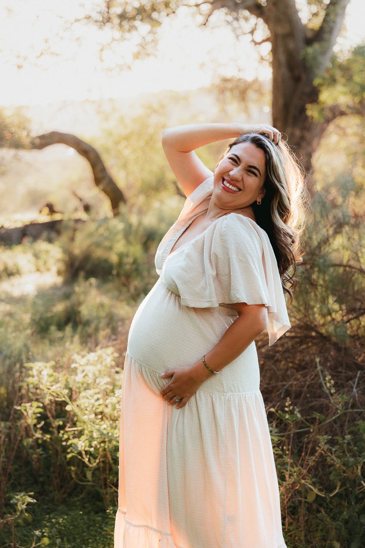 Expecting mom in a flowing peach and cream dress laughing freely during an outdoor maternity session at golden hour, captured in warm natural light by an Orange County maternity photographer.