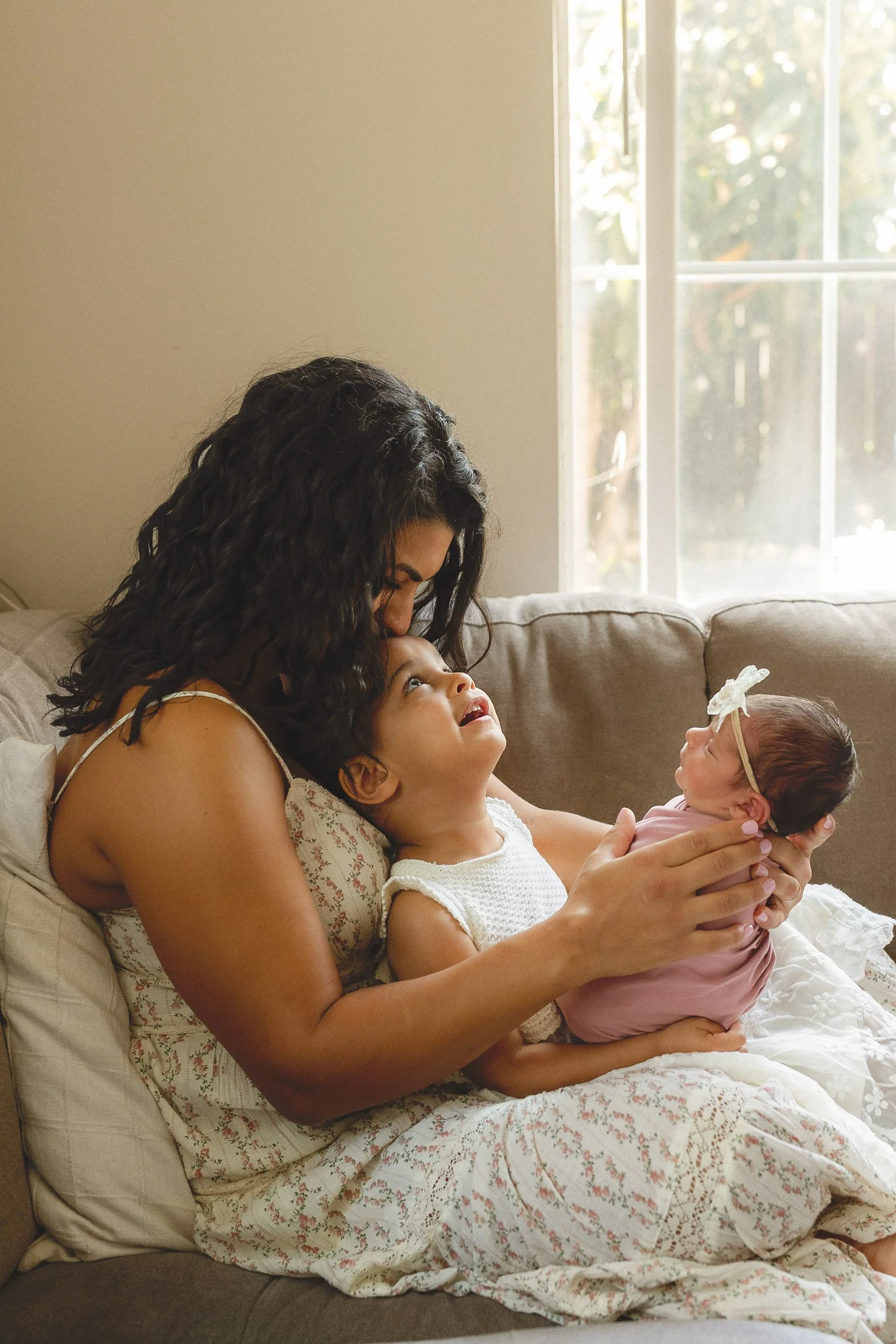 Mom sitting on a couch with her toddler while holding their newborn in soft window light, Orange County photographer.