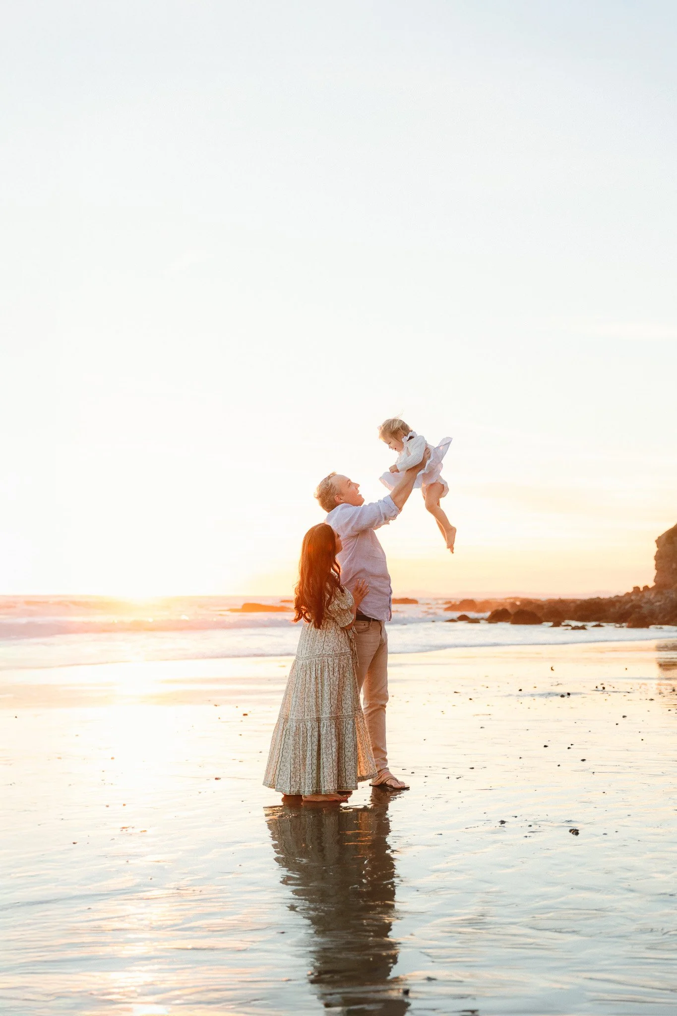 Orange County family photographer capturing a parent lifting their baby at sunset on the beach while their partner stands close.