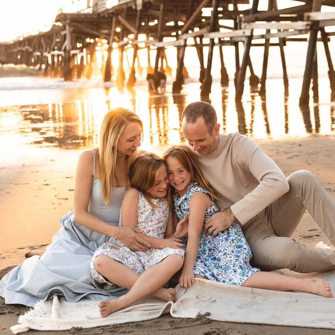 Family sitting on a blanket on the beach with the San Clemente Pier pilings and sunset light behind them.