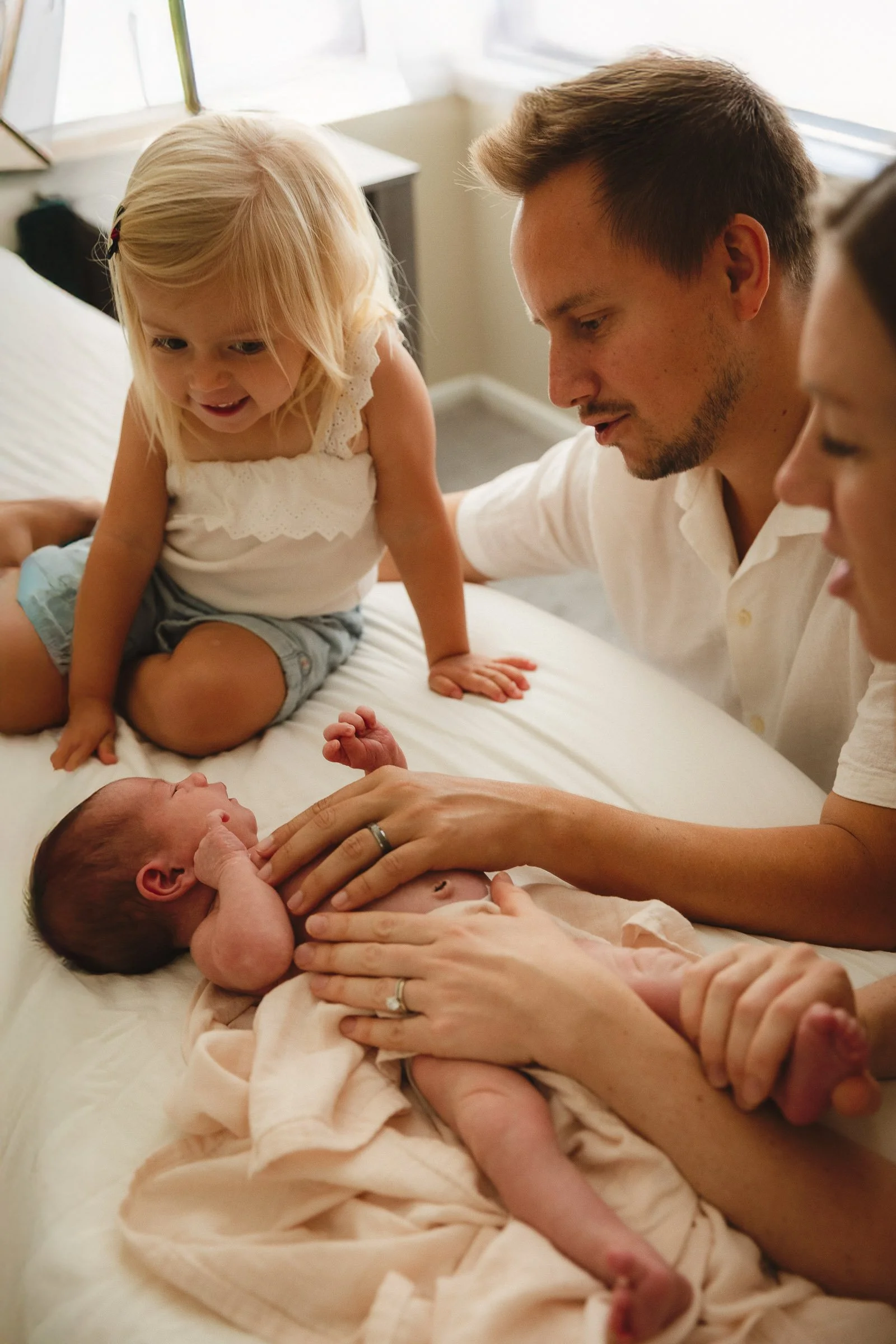 Toddler meeting a newborn on the bed while parents gently comfort baby during an in-home session with an Orange County newborn photographer.