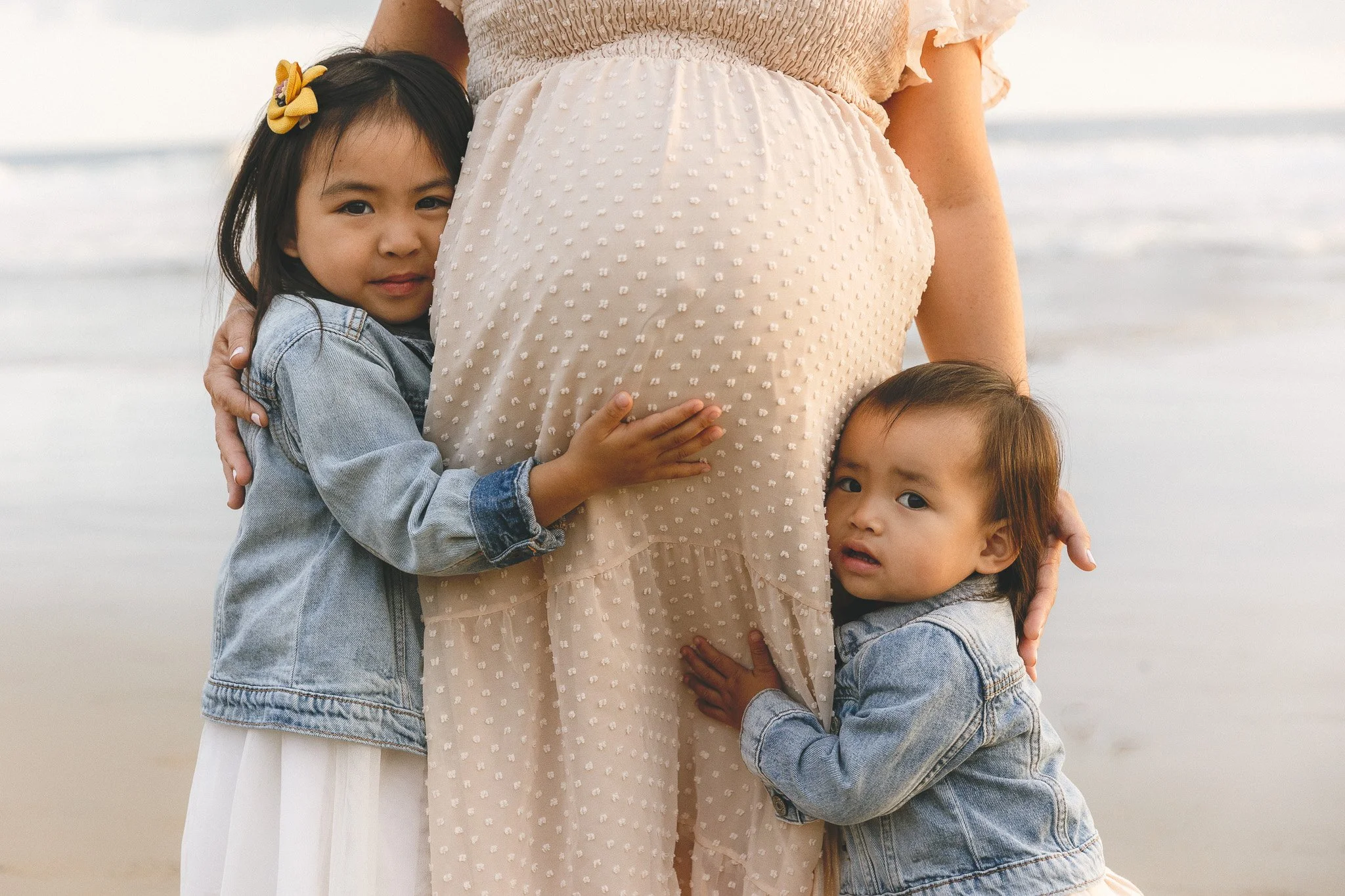 Two siblings in denim jackets hugging their pregnant mom's belly during a beach maternity session in Orange County