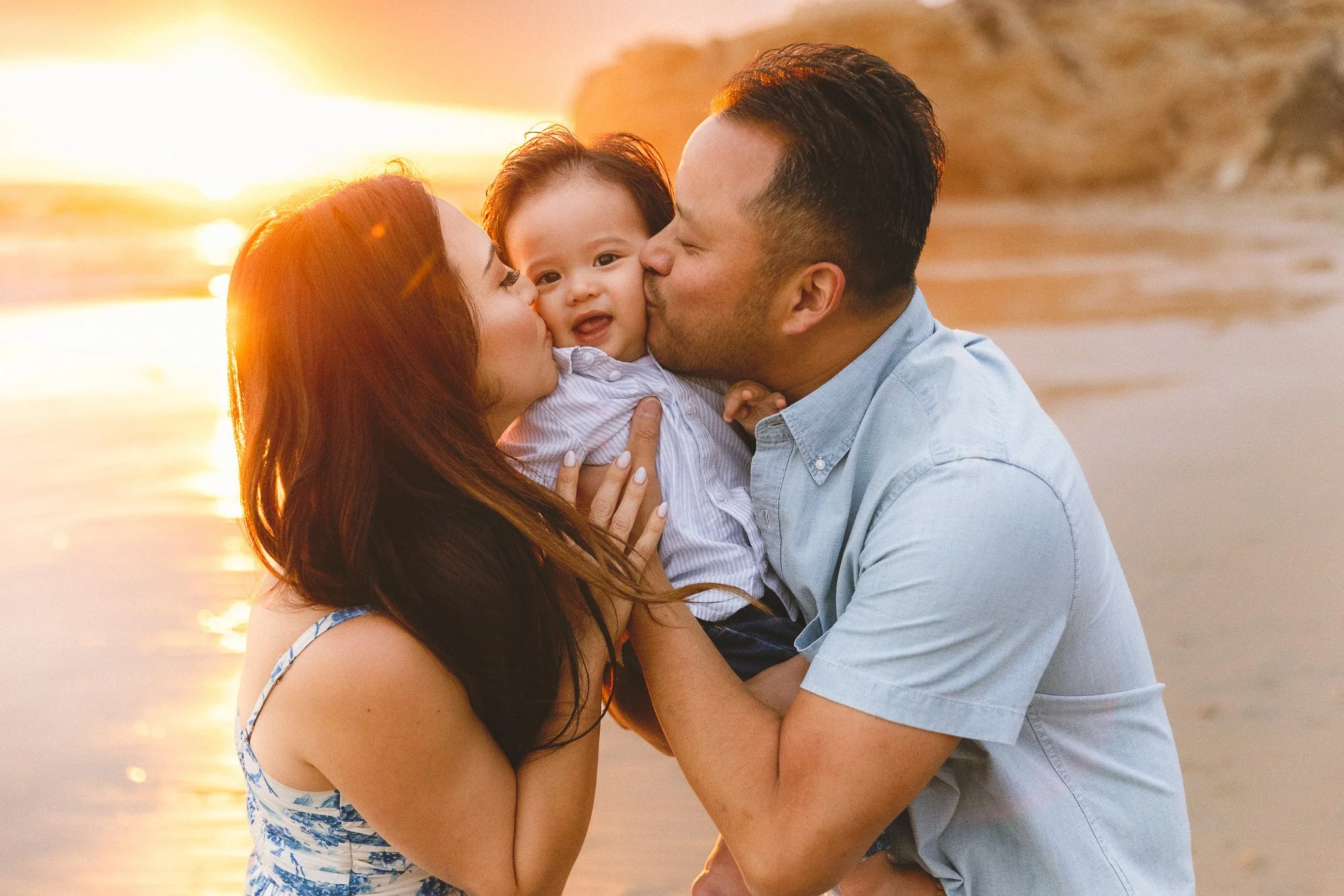 Orange County family photographer capturing parents kissing their toddler’s cheeks at sunset on the beach.