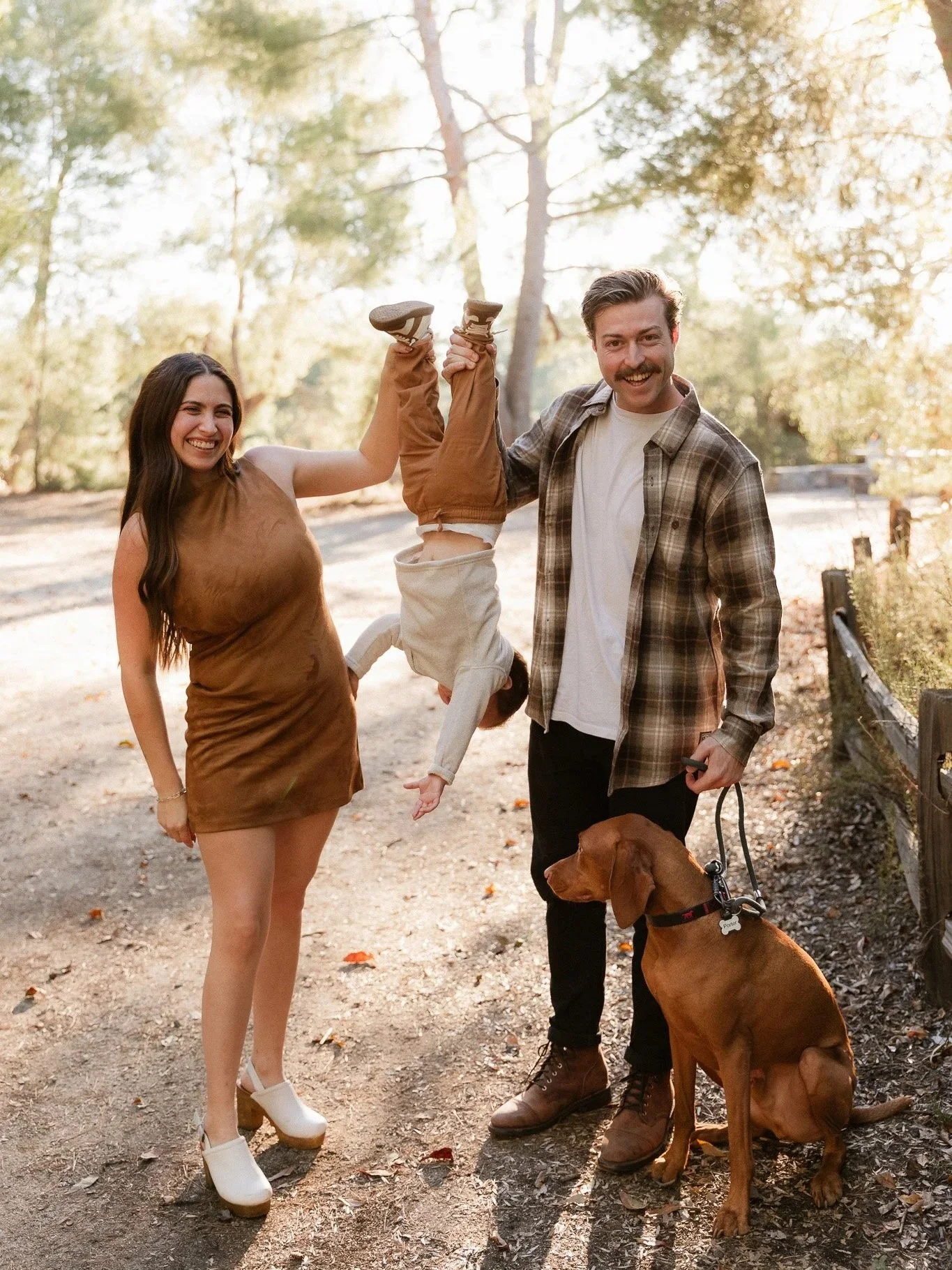 Parents laughing while their toddler hangs upside down on a sunny trail, with their dog beside them.