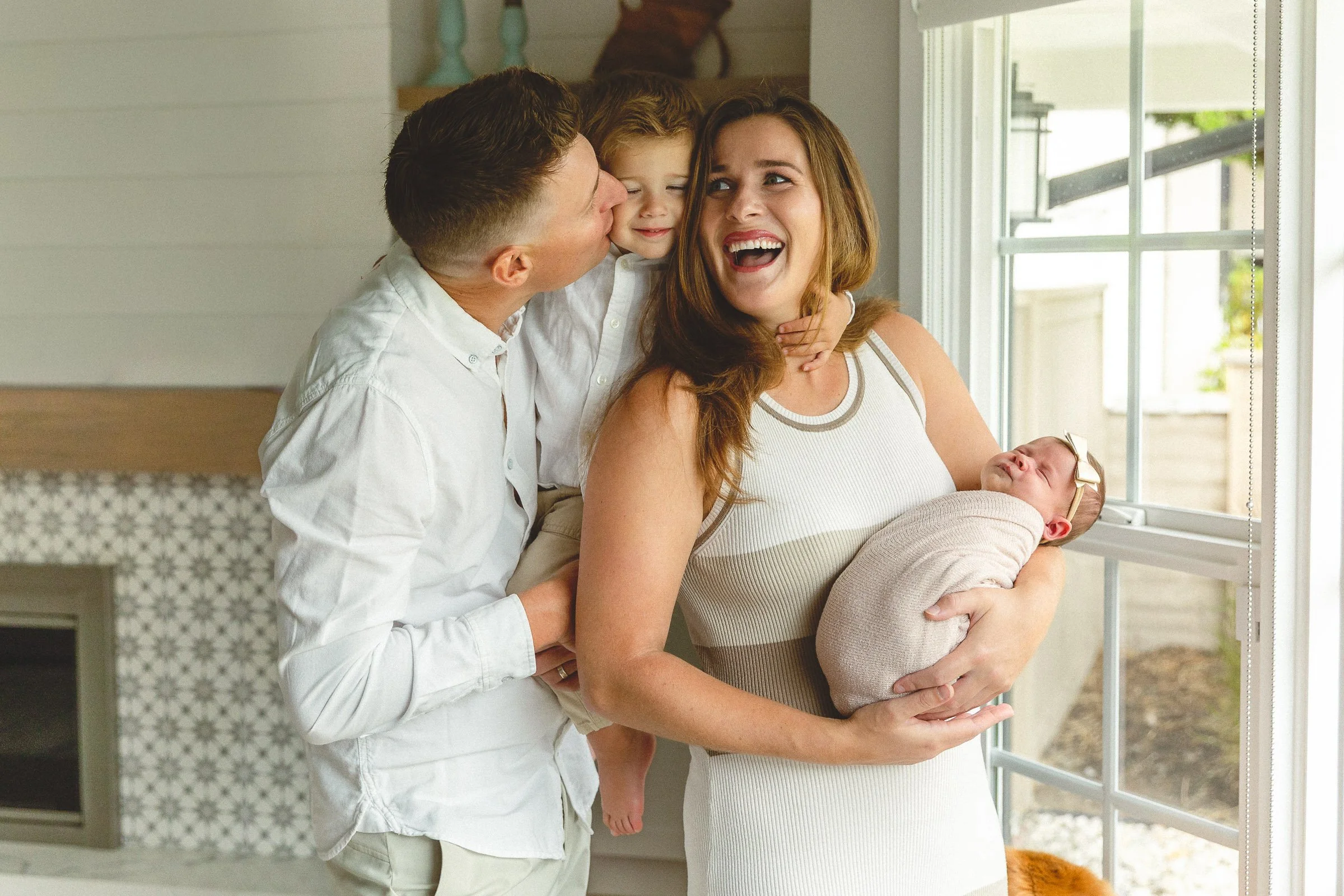 Family of four laughing together as dad holds toddler and mom cradles their newborn by a window during an in-home newborn session