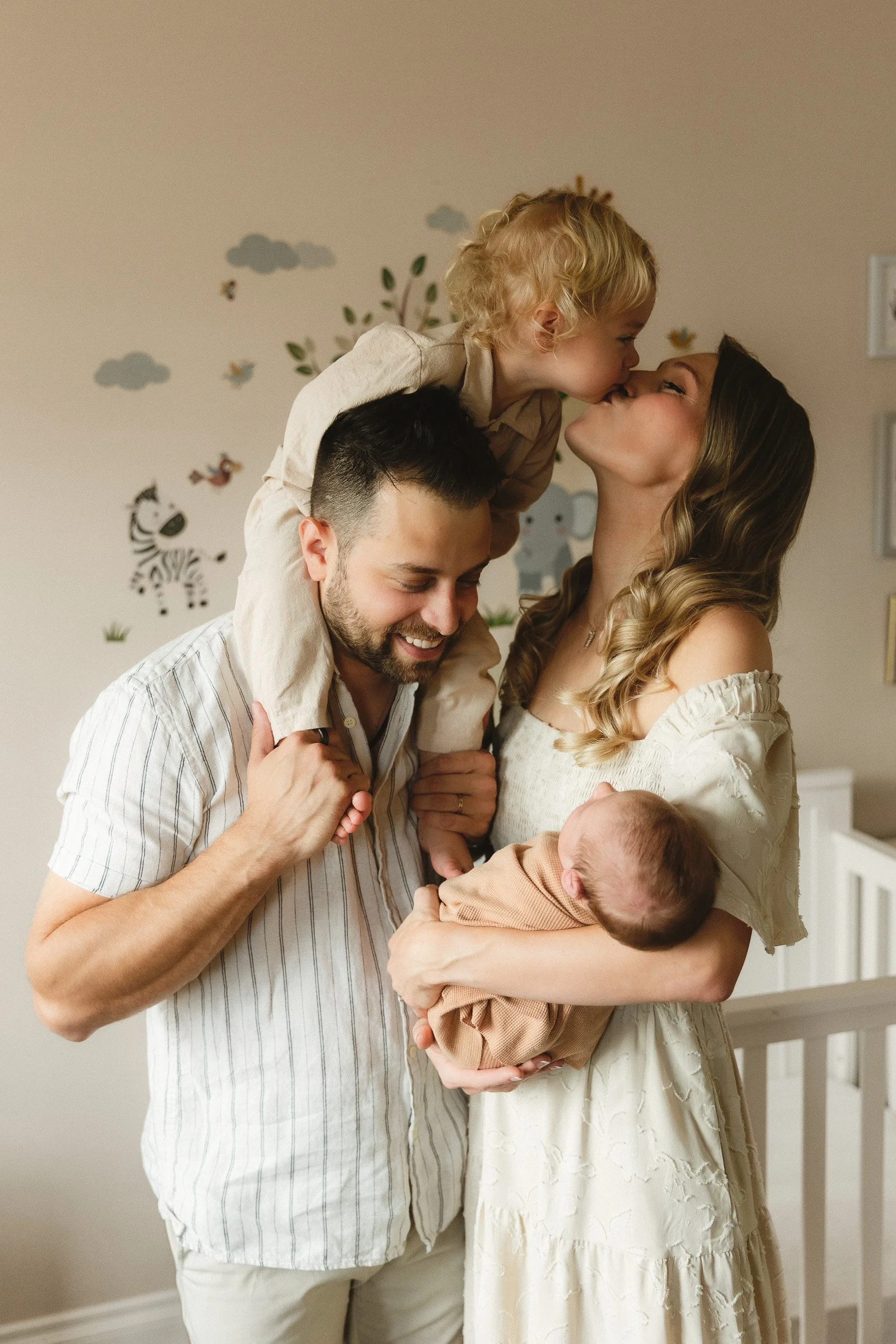 In-home newborn photography session with an older sibling in Orange County. Dad holds his toddler on his shoulder while mom cradles the newborn in a soft wrap, all standing in the nursery.