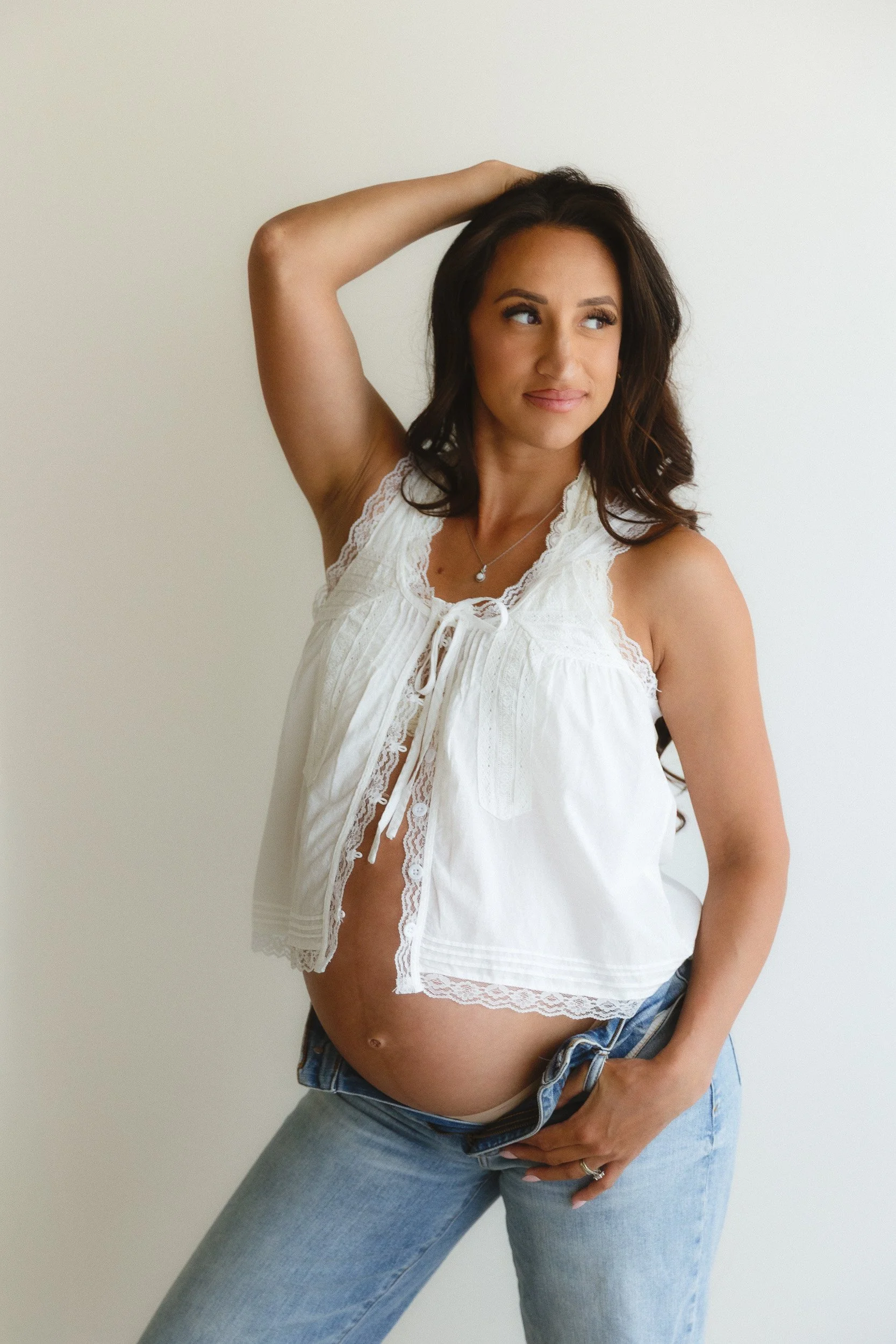 Expecting mom in a white lace top and unbuttoned jeans standing in soft natural window light during a studio maternity session, captured by an Orange County maternity photographer in Huntington Beach.