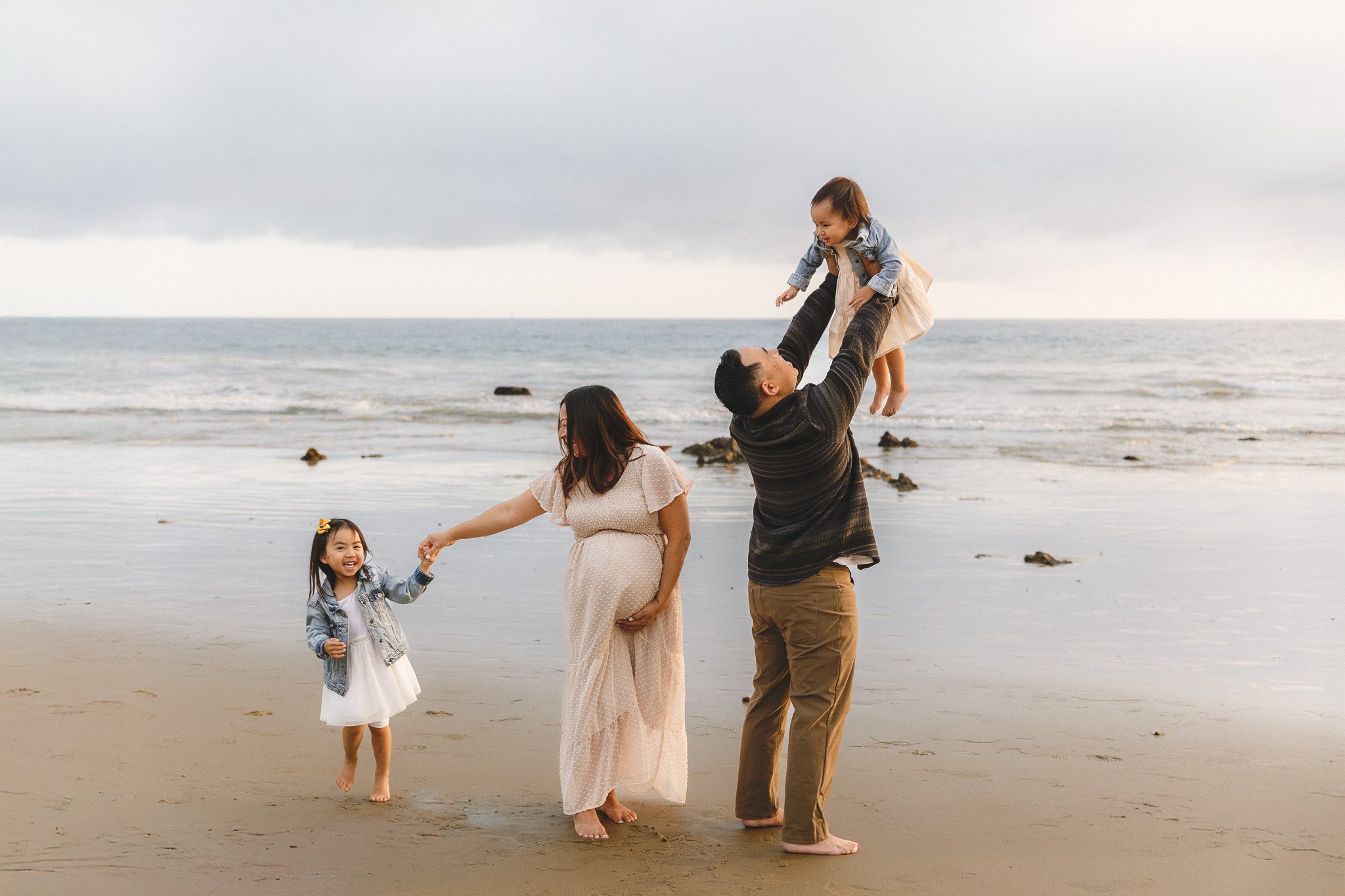Maternity family photos with a toddler at an Orange County beach. Pregnant mom holds her older daughter's hand while dad lifts their younger daughter into the air.