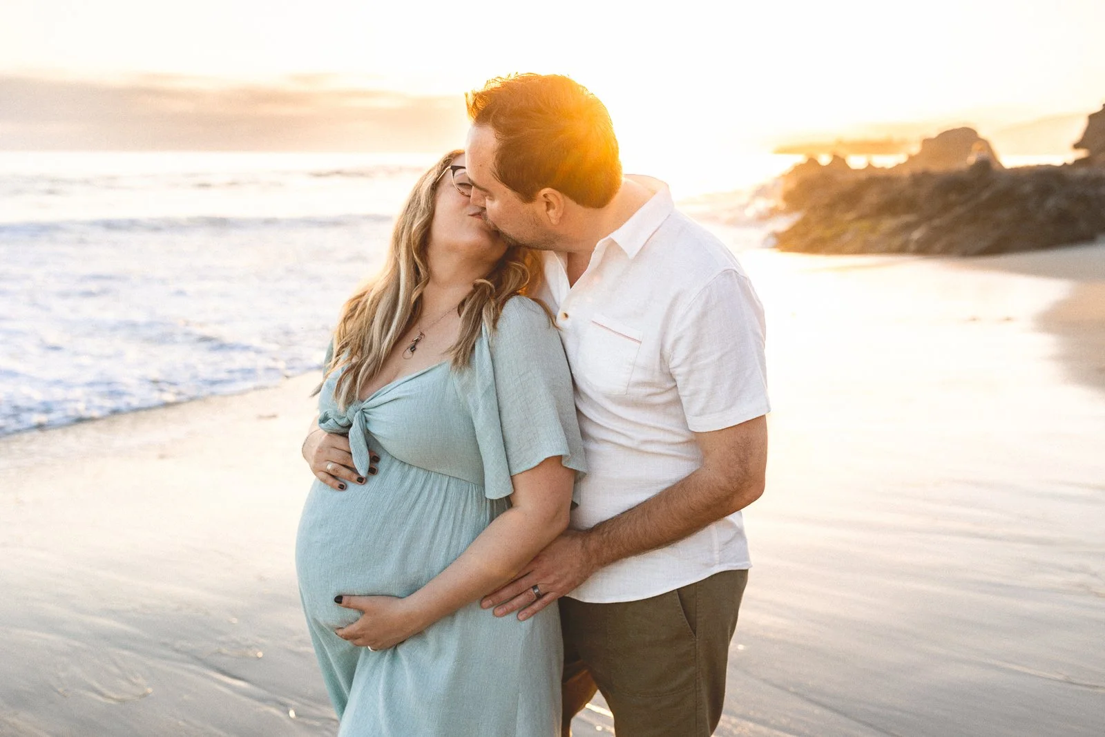 Couple kissing at golden hour while mom cradles her pregnant belly during a third trimester maternity session in Orange County
