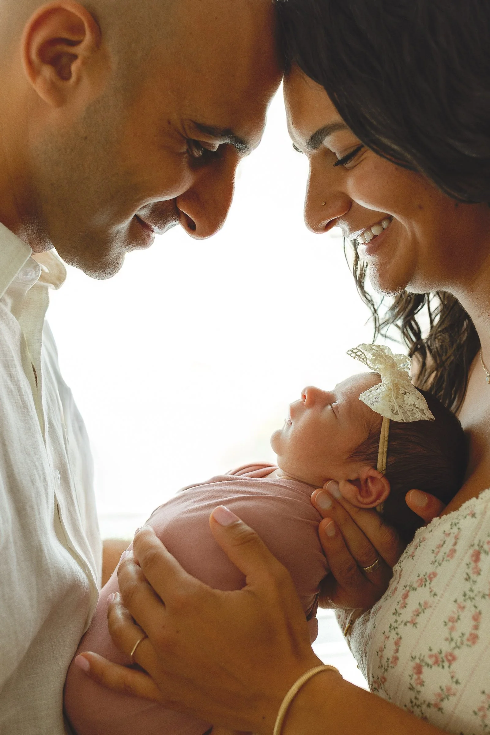 Parents leaning in close, smiling while holding their newborn in bright window light, Orange County newborn photographer.