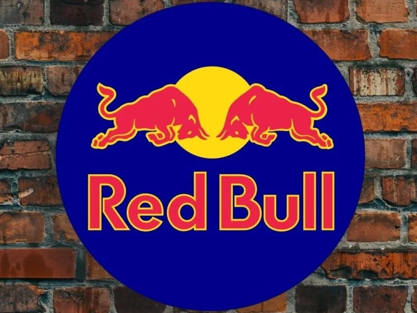 Red Bull logo with two red bulls facing each other in front of a yellow circle, on a blue circular background, mounted on a brick wall.