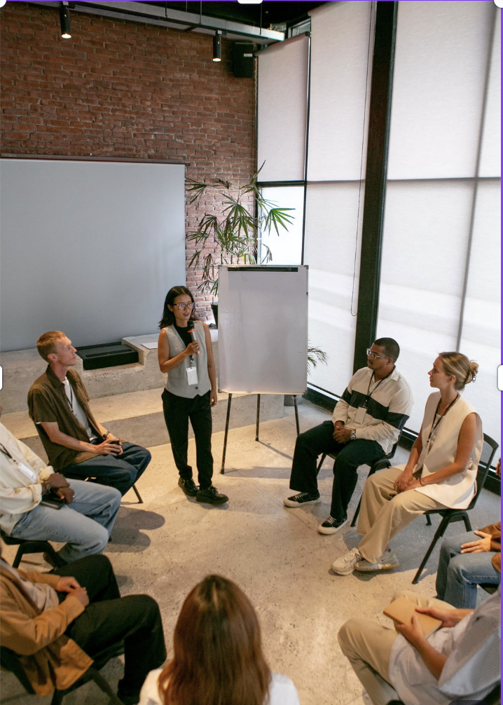 A woman standing and speaking into a microphone in front of a flip chart, with five seated people listening in a modern room with brick walls and large windows.