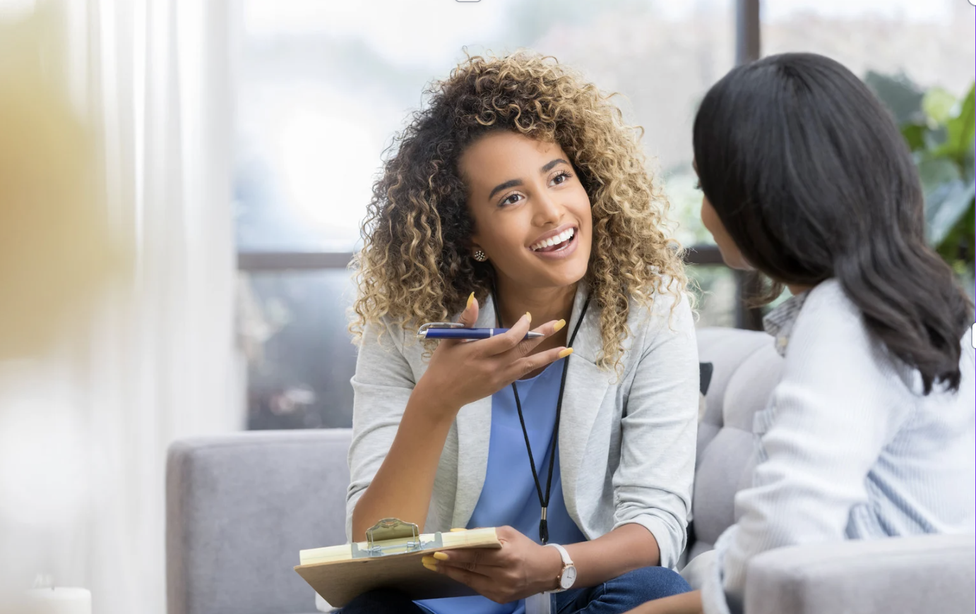 Two women having a conversation in a bright, modern office or lounge with large windows and natural light.