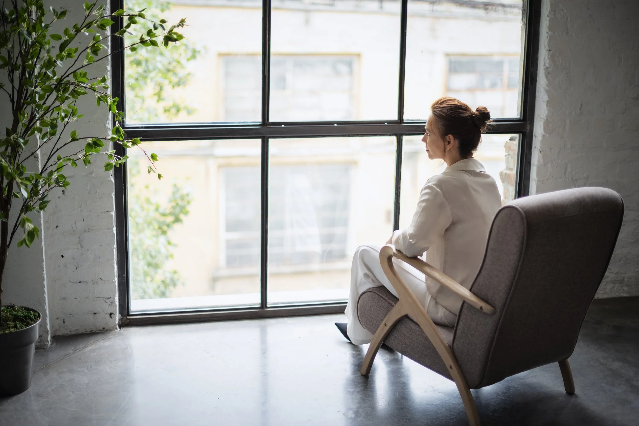 A woman with brown hair tied back, sitting comfortably in a modern gray armchair near a large window with black metal framing. She appears to be relaxed, gazing outside. There is a tall green plant in a pot on the left side of the image, and the room has a white brick wall. Outside, neighboring buildings and trees are visible through the window.