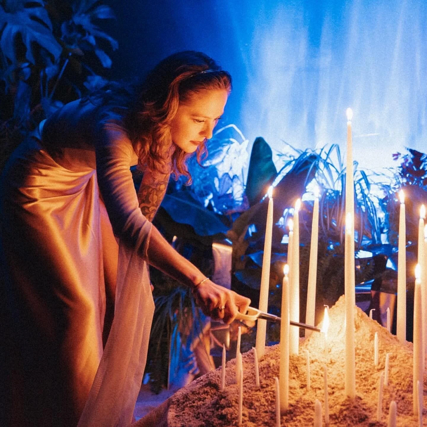 A woman with tattoos on her right arm, wearing a pink dress, lighting a candle at a large display of lit candles on pink sand, with tropical plants and blue lighting in the background.