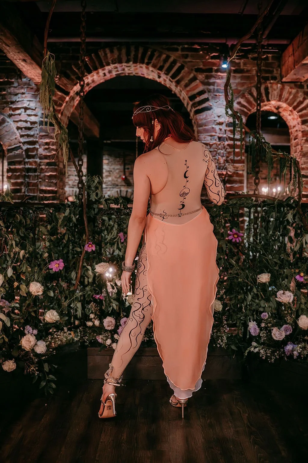 A woman stands with her back to the camera in a dimly lit, rustic interior with exposed brick walls and floral decorations. She is wearing a peach-colored, sheer skirt and high heels, with tattoos and jewelry, and a headband.