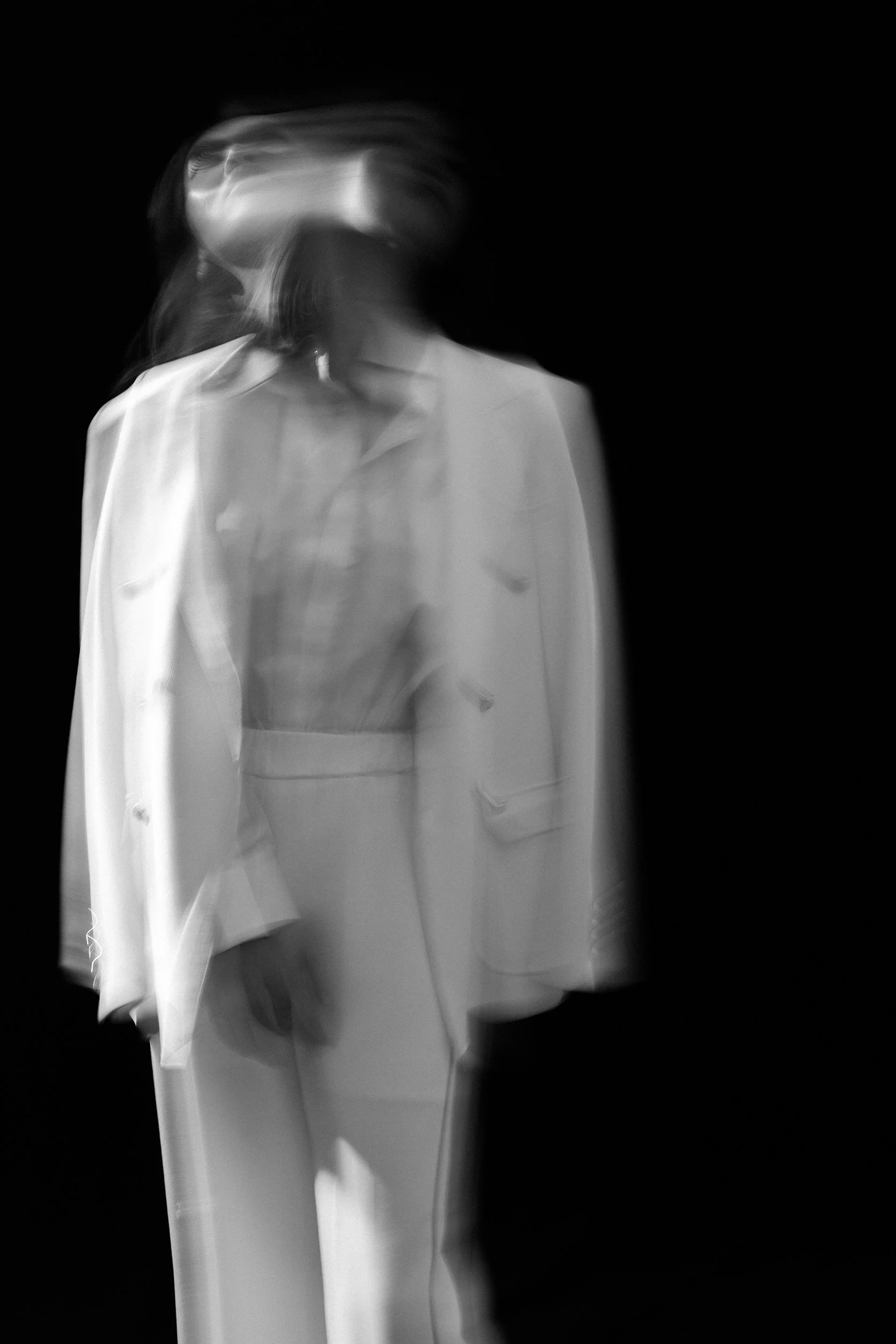 Black and white photo of a woman wearing a white blazer, with motion blur. The background is solid black.