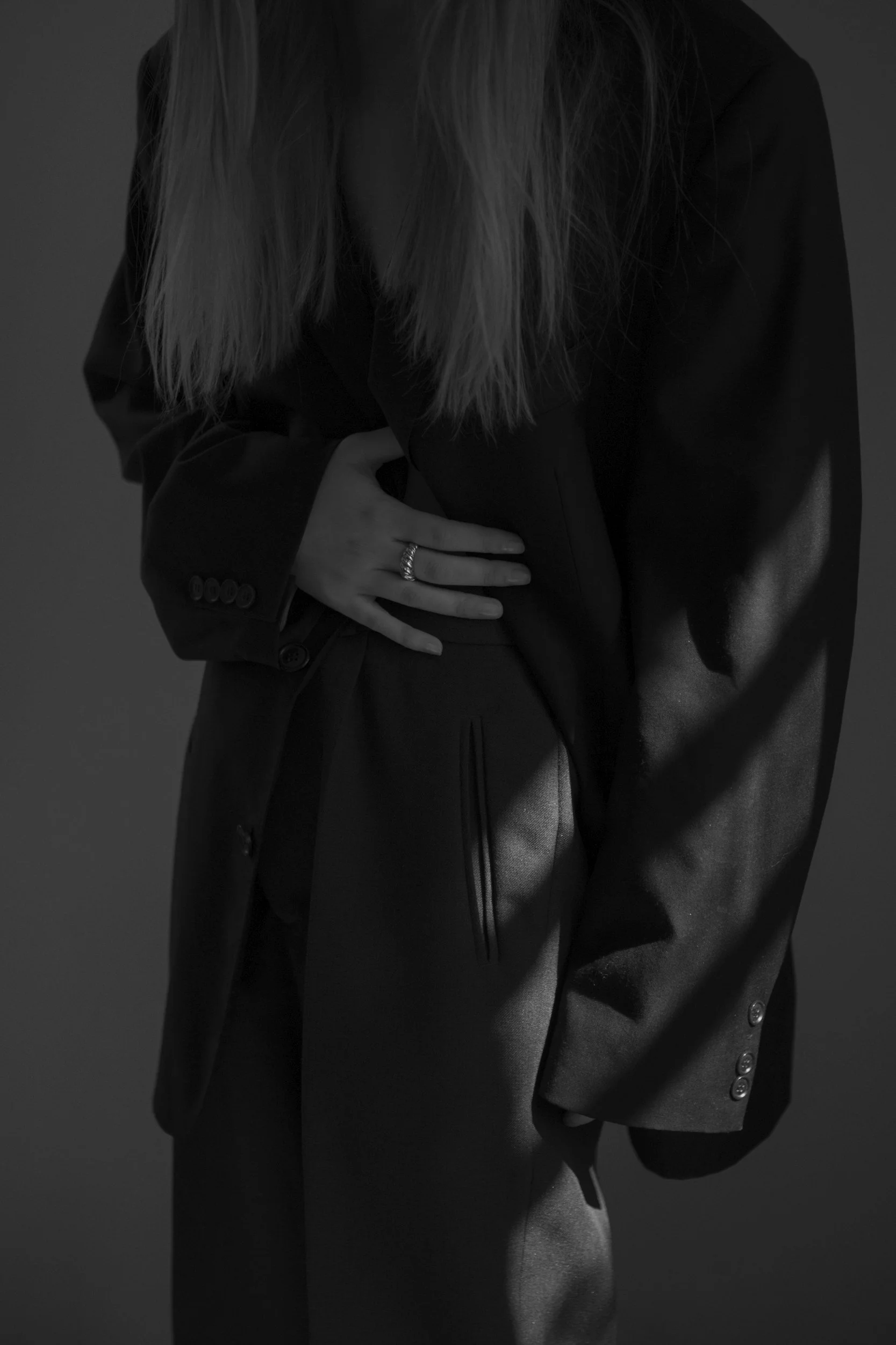 The image shows a person wearing a black blazer with button details on the sleeve and grey pants. The person's hand, with rings on the fingers, is resting on their waist. The person's face is not visible, and the photo is in black and white with a dark background.