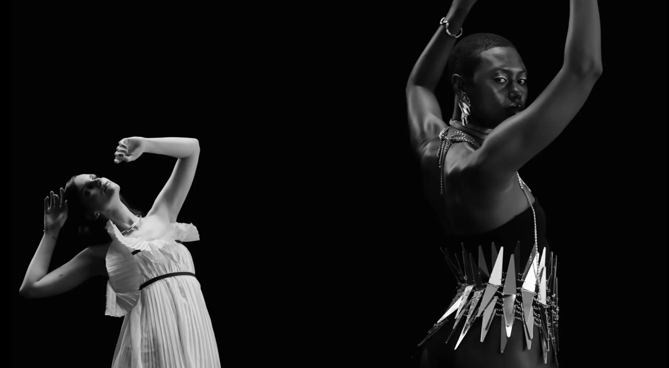 Black and white photo of two dancers, one in a white dress with ruffles, and the other in a dark, embellished outfit, posing artistically against a black background.