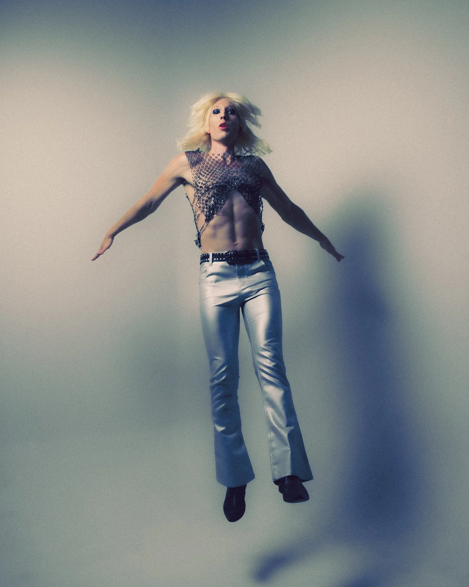 A person with blonde hair and makeup in a fashion pose, wearing a revealing black mesh top and silver pants, standing against a plain background.
