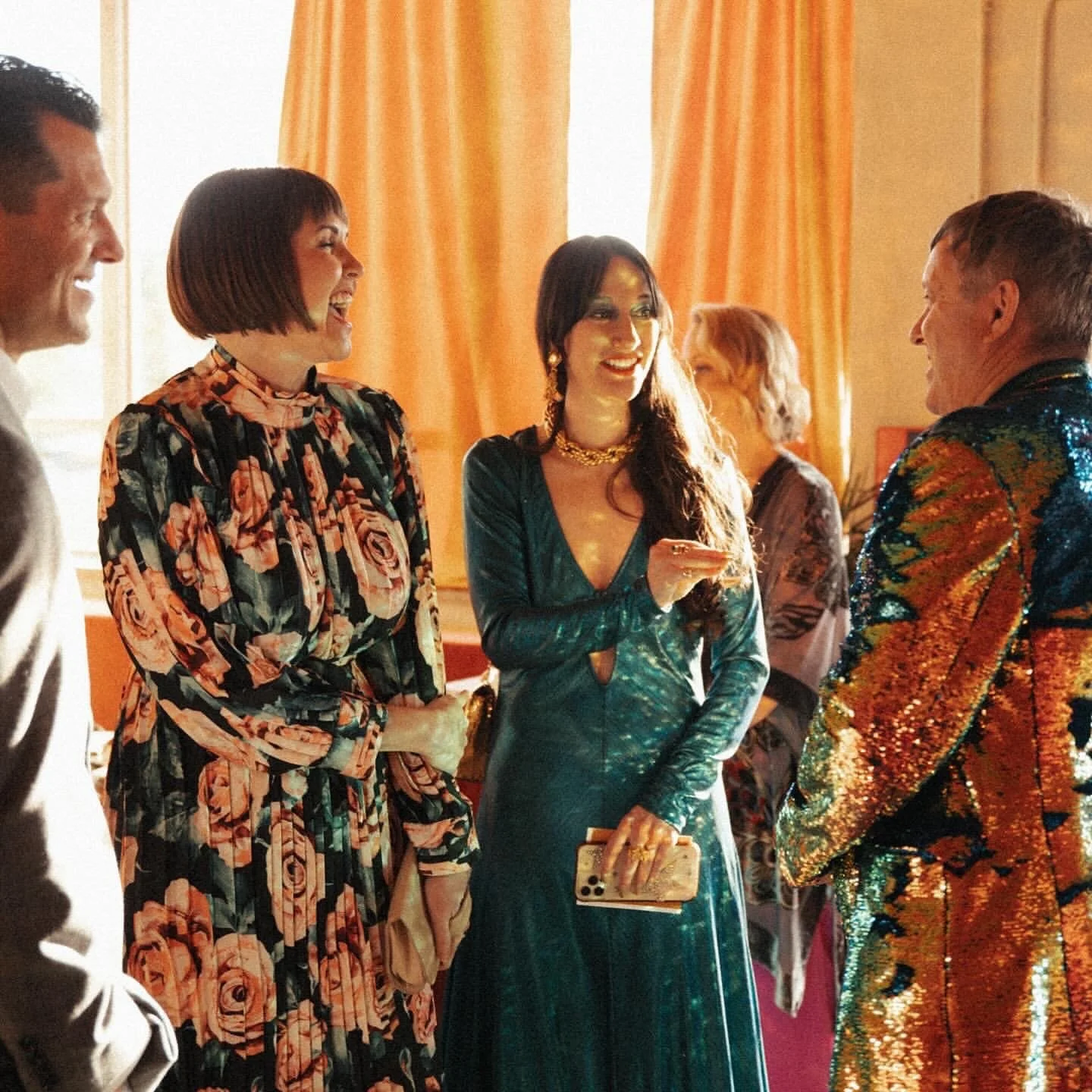 Group of people at a social gathering, dressed in colorful and fashionable clothing, engaging in conversation and smiling.