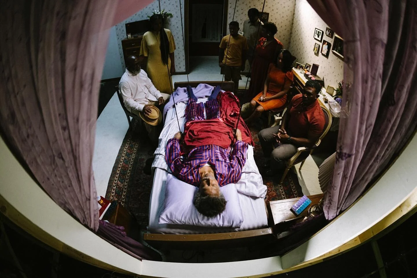 People gathering around a person lying in bed, looking at them through a window or from above, in a room decorated with family photos and furnishings.