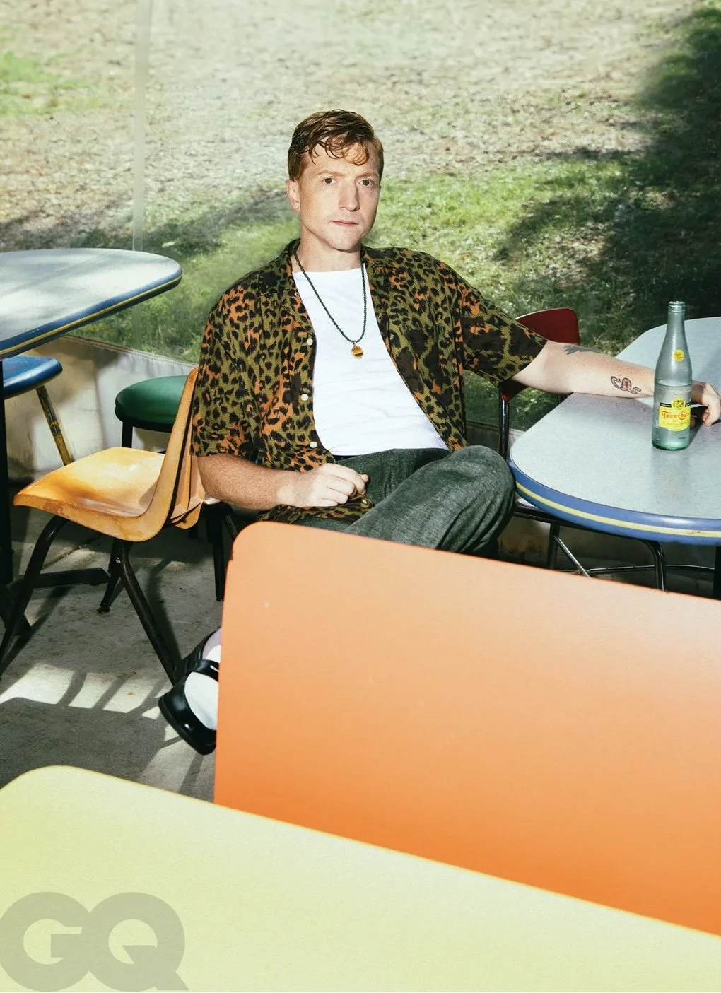 A man with short brown hair sitting at a colorful table in front of a backdrop of a nature scene, holding a green beverage bottle. He is wearing a leopard print shirt over a white T-shirt and has a tattoo on his left arm.
