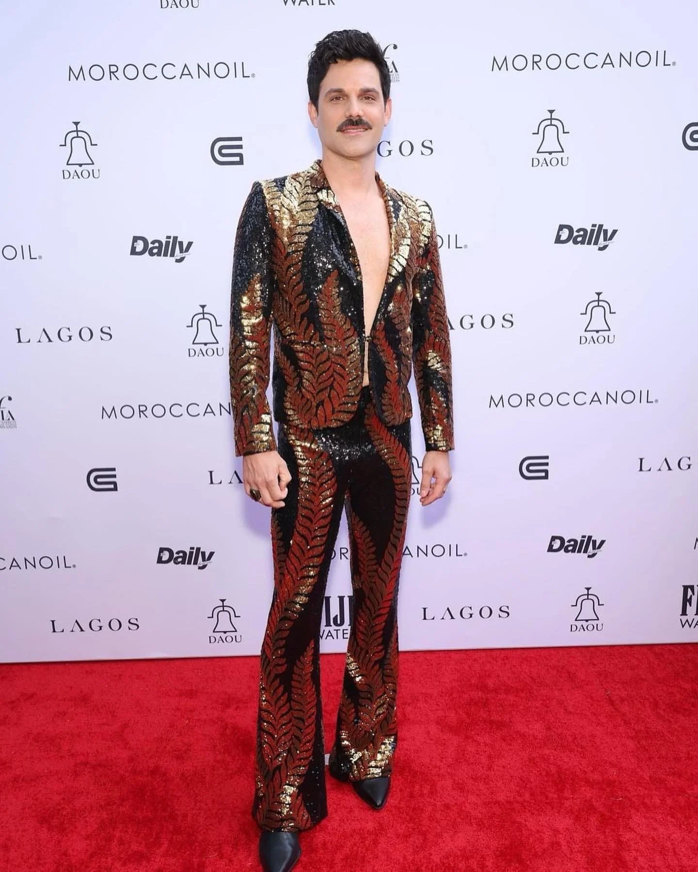 Man in a black, gold, and red patterned embroidered suit with a deep V-neckline, standing on a red carpet in front of a white backdrop with logos and text.