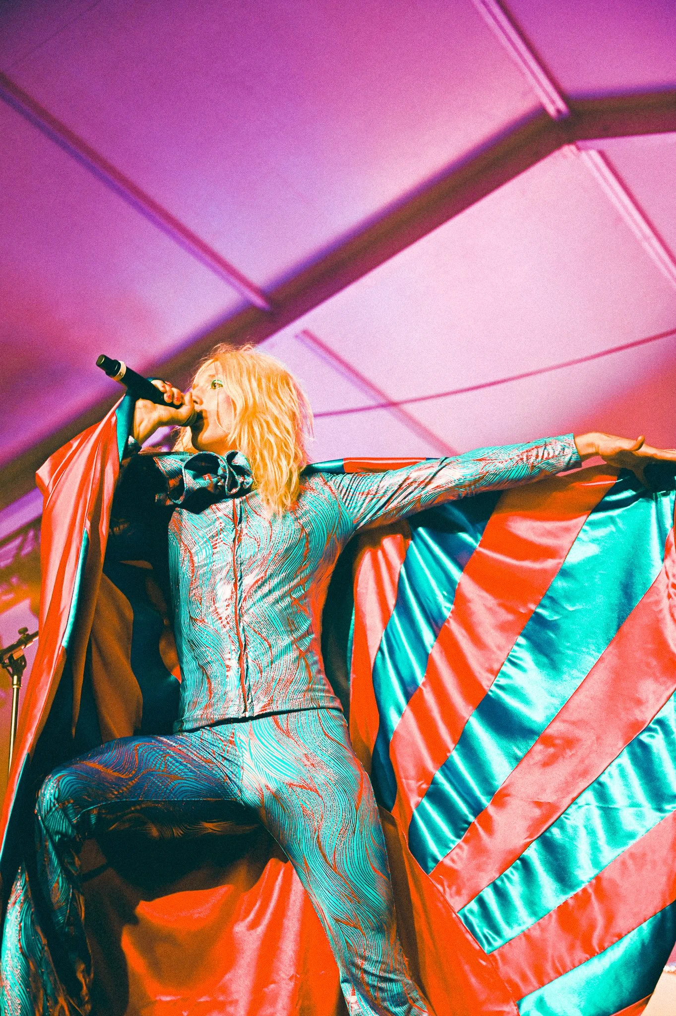 A performer with blonde hair singing into a microphone on stage, wearing a colorful patterned outfit and a red, teal, and orange cape, with a pink and purple illuminated ceiling in the background.