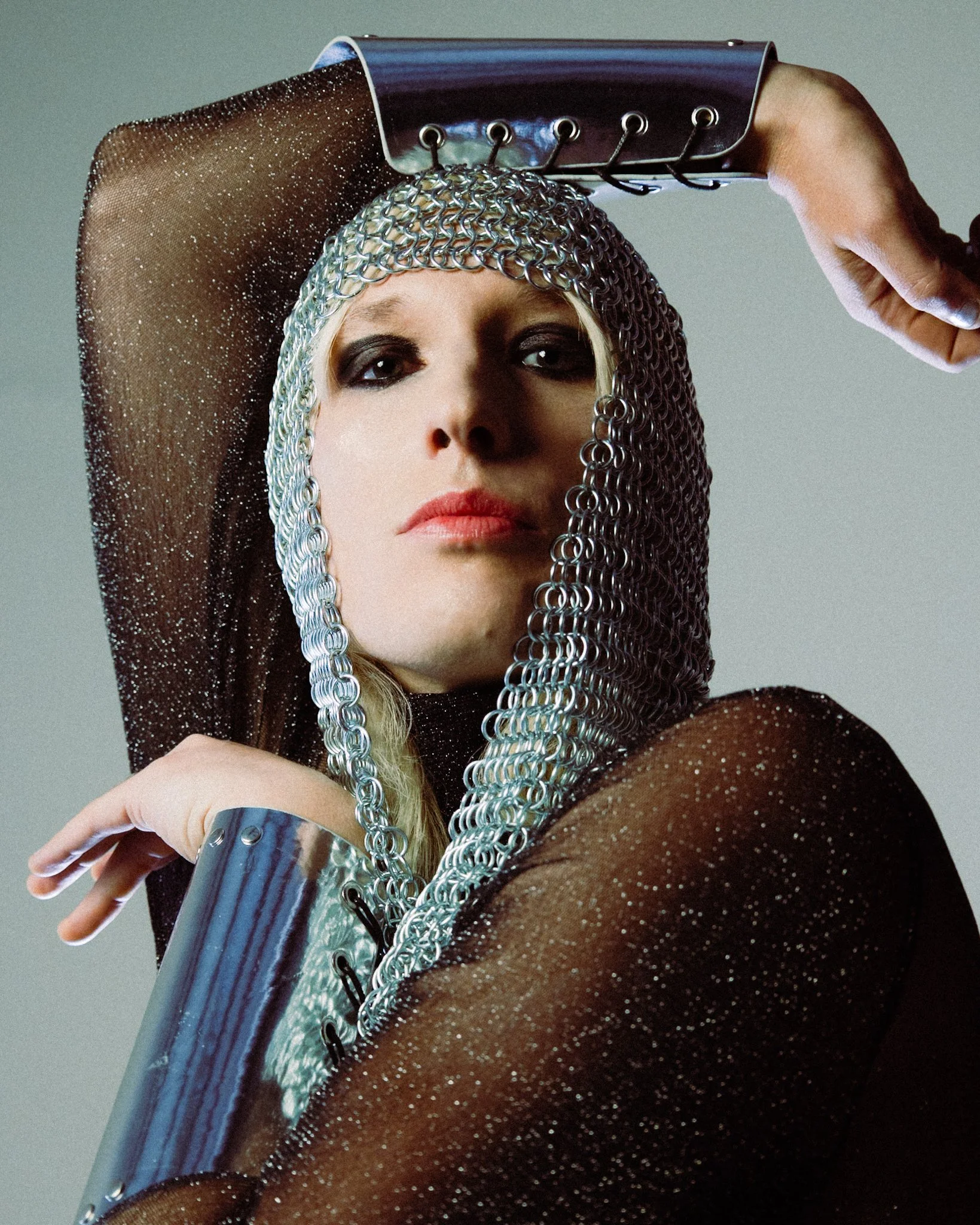 A woman with blonde hair and dark eye makeup is wearing a chainmail hood and a black sheer top with glitter. She is holding a metallic object above her head with her right hand, with her left hand resting on her upper arm.