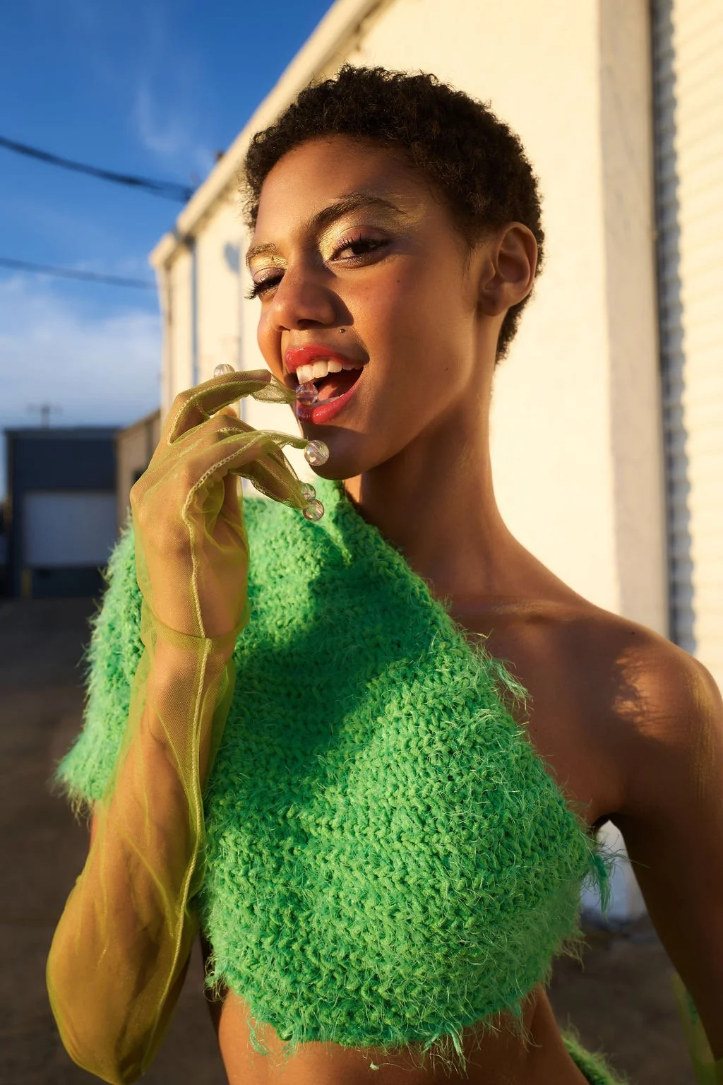 A woman with short curly hair posing outdoors in bright sunlight, wearing a textured green top and sheer yellow gloves, holding a pearl necklace near her mouth.