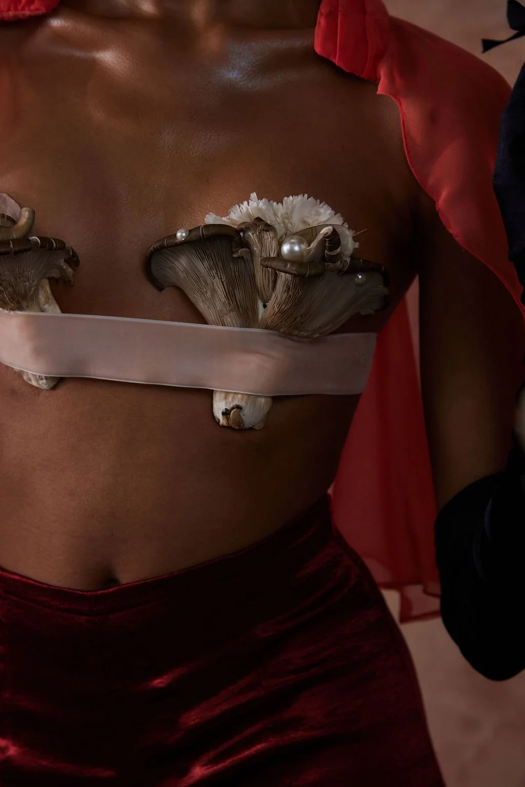Close-up of a woman's torso with a creative top made of mushrooms, pearls, and flowers, paired with red satin pants and a red sheer shawl.