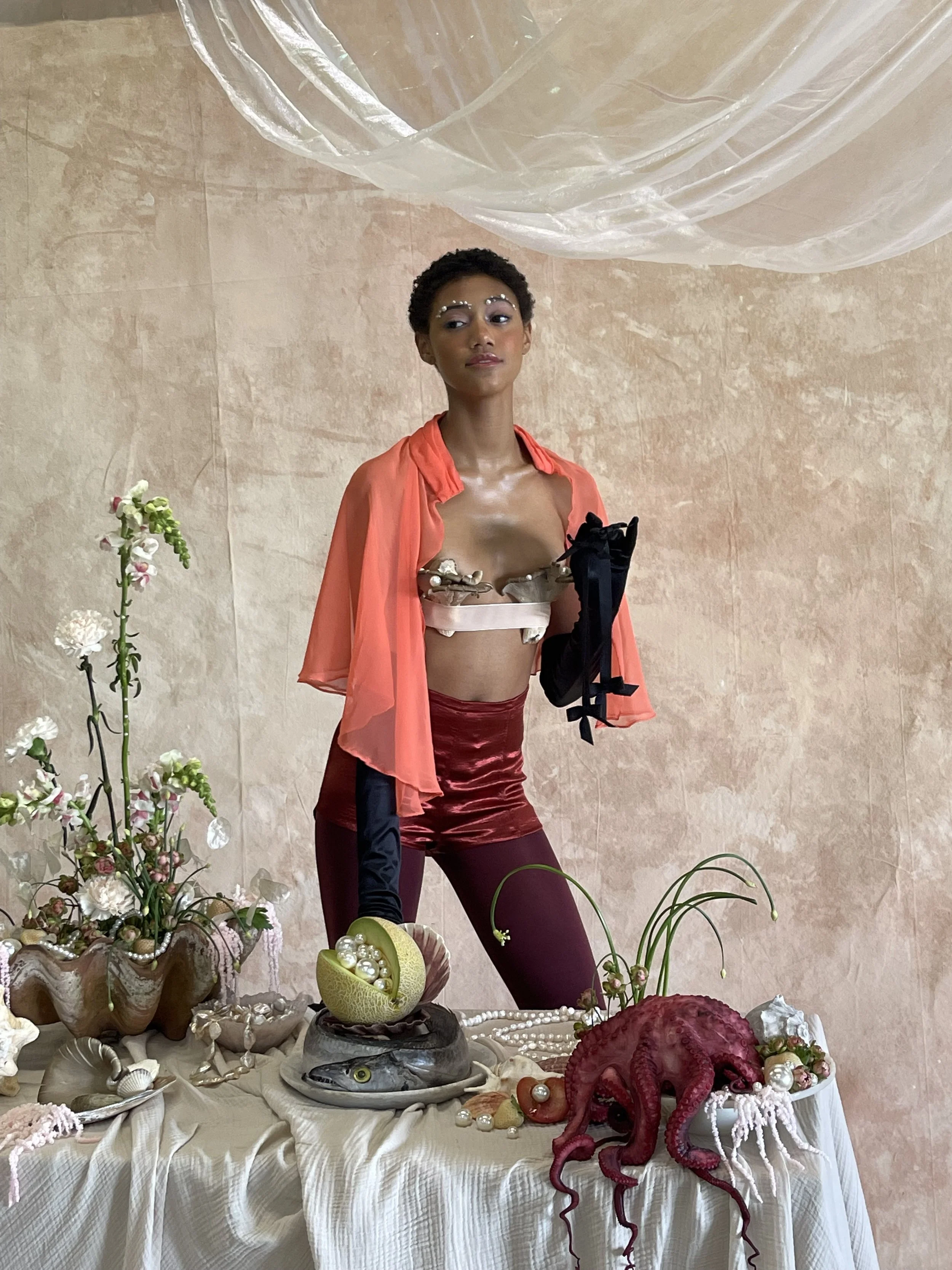 A woman dressed in artistic fashion with makeup and jewelry, standing behind a table decorated with flowers, seashells, a fish, and an octopus, against a pinkish textured background.