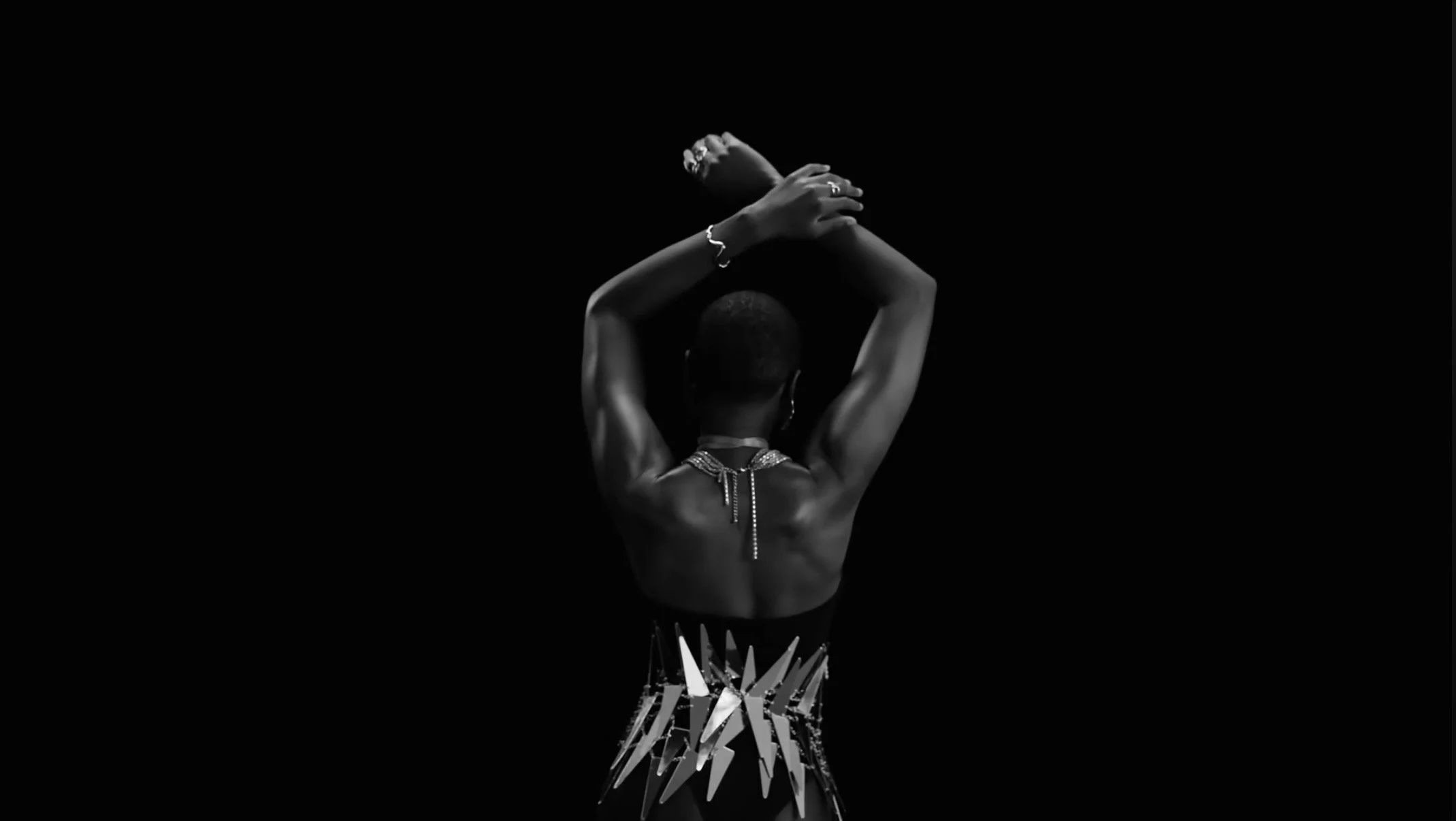 Black and white photograph of a person from behind with their hands crossed over their head, adorned with jewelry, wearing patterned clothing, and standing against a dark background.