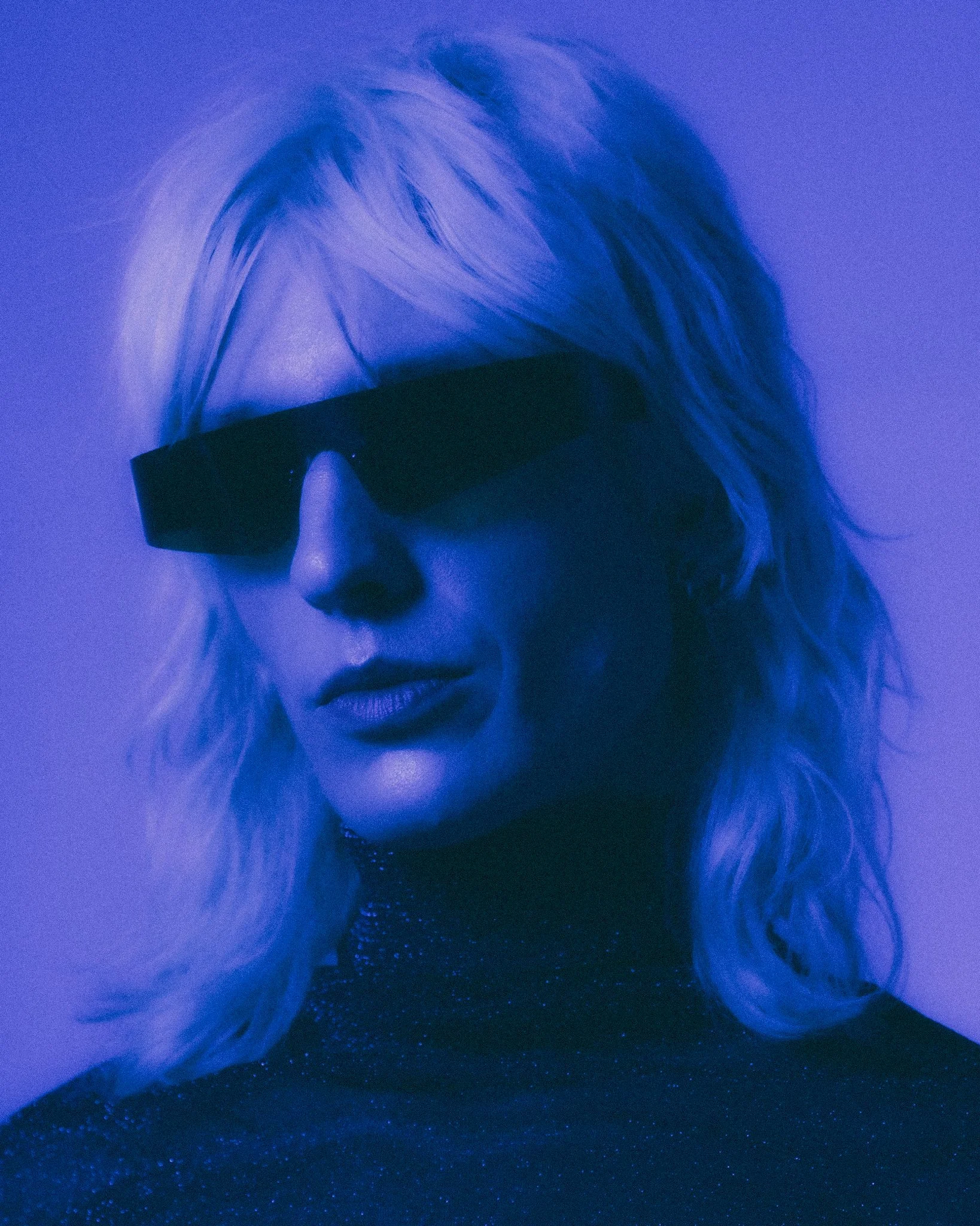 Close-up of a woman's face with platinum blonde hair, wearing black sunglasses, a choker necklace, and a black top, illuminated with blue lighting.