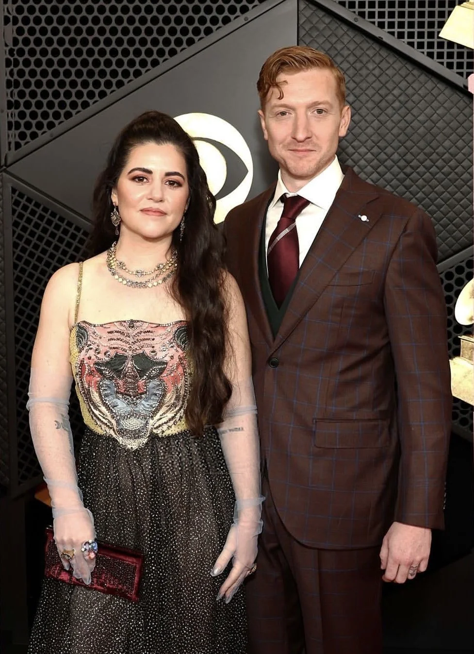 A man and woman dressed in formal attire standing together in front of a black textured backdrop with a white logo.
