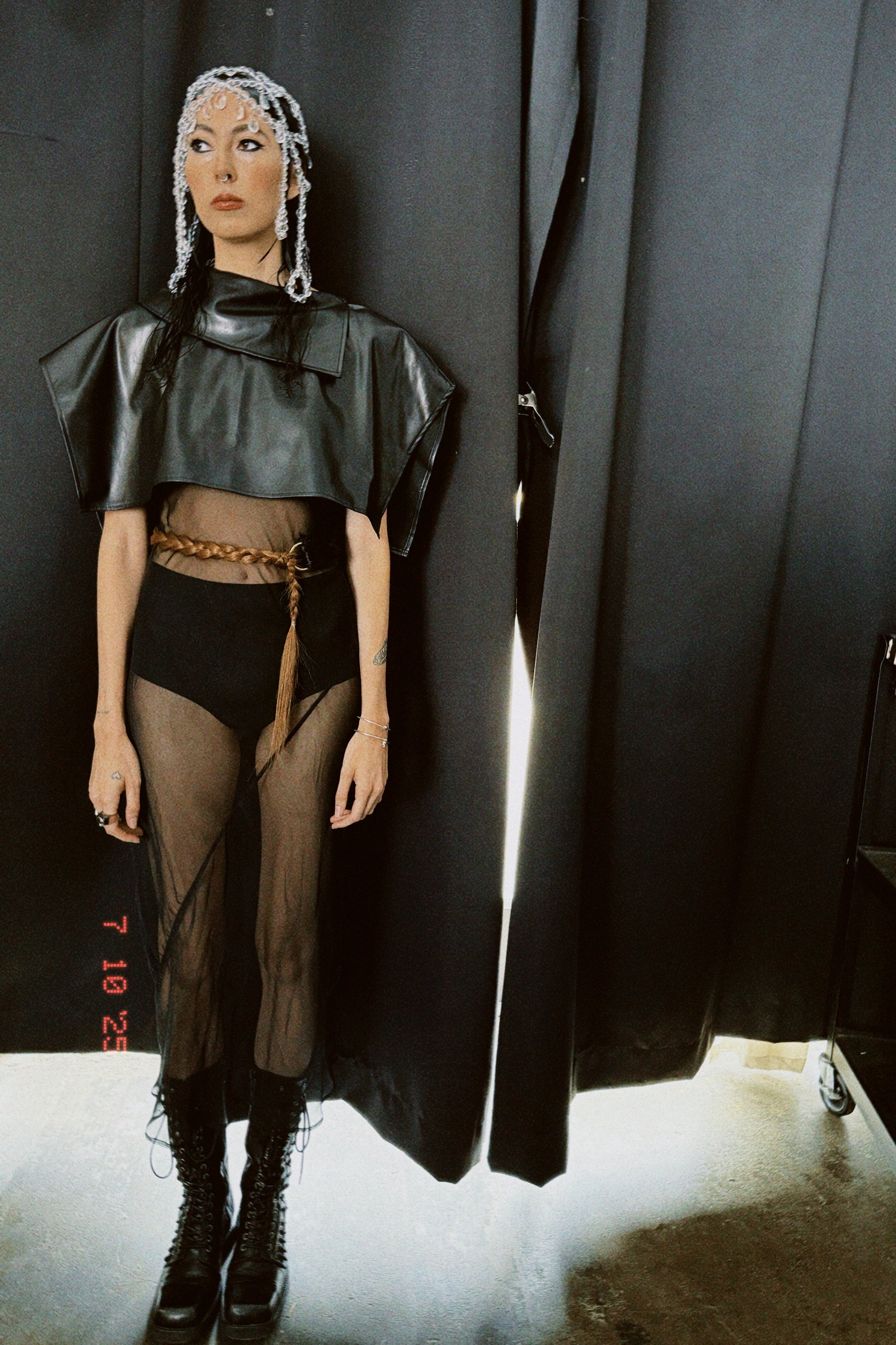 A woman with long dark hair and wearing dark eye makeup stands in an elevator with black panels. She has a nose ring and tattoos on her left arm. She is dressed in a sheer black skirt, black boots, and a cropped leather jacket draped over her shoulde