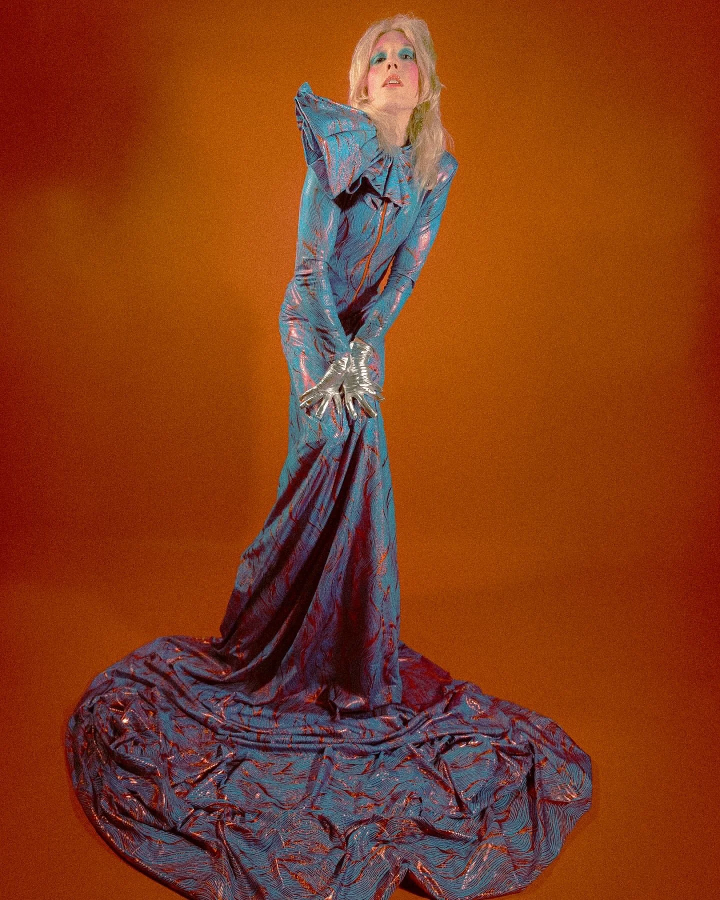 A woman with platinum blonde hair, sporting blue eyeshadow and pink lipstick, wearing a shiny, patterned blue and red gown with large, exaggerated ruffled shoulders and a long train, stands against an orange background.