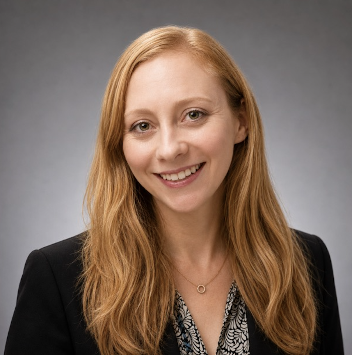 Headshot of a woman with long red hair, smiling, wearing a black blazer and patterned blouse, against a gray background.