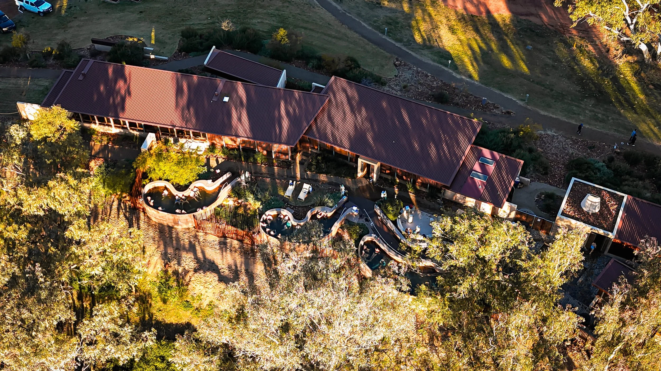 An aerial view of a modern building with a red roof, surrounded by trees and landscaped outdoor areas including a winding water feature and seating areas. There are a few people walking nearby.