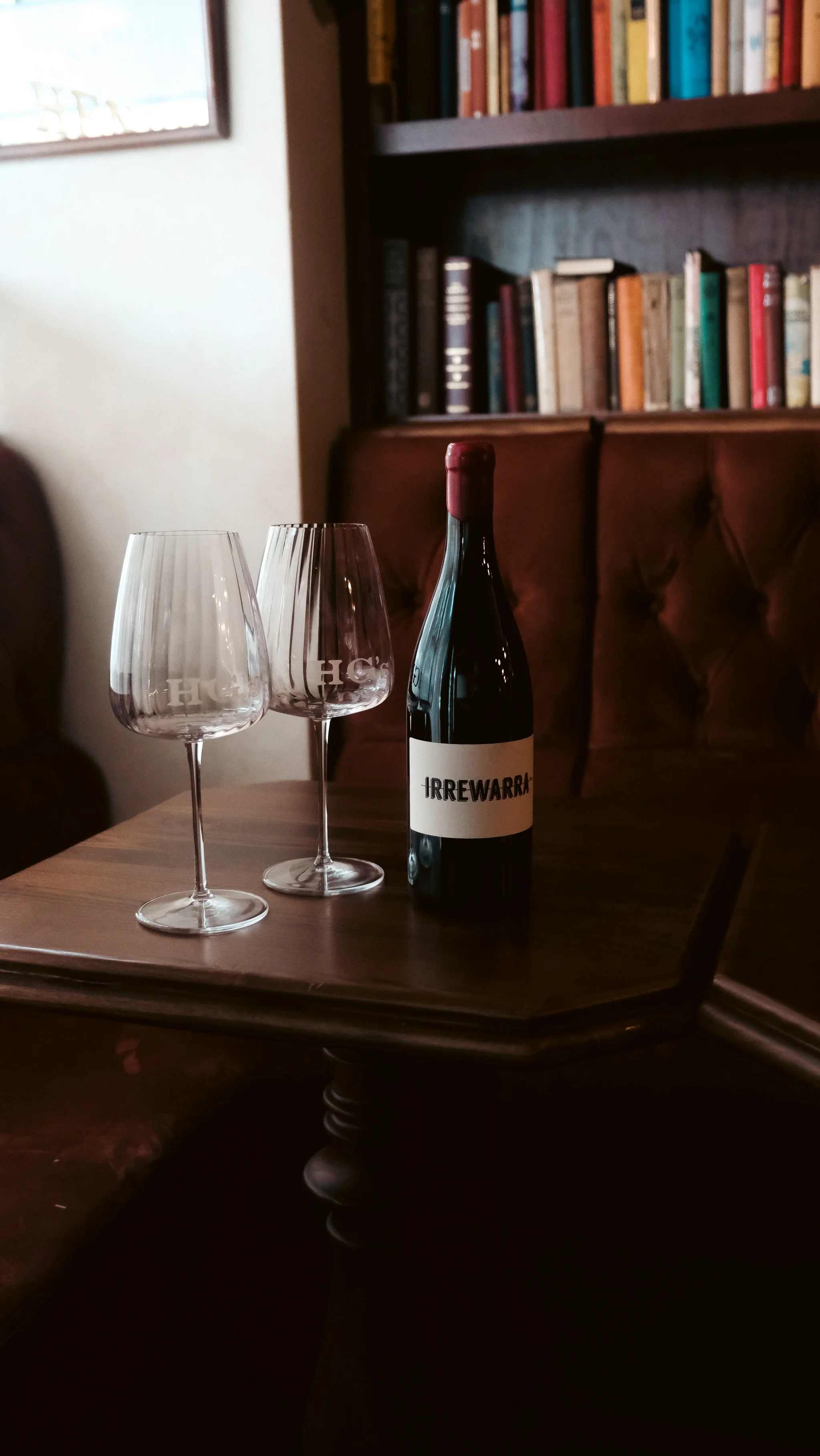 A bottle of red wine labeled 'IRREWARRI,' two empty wine glasses with the letters 'HCS' etched on them, placed on a wooden table in a cozy room with a bookshelf in the background.
