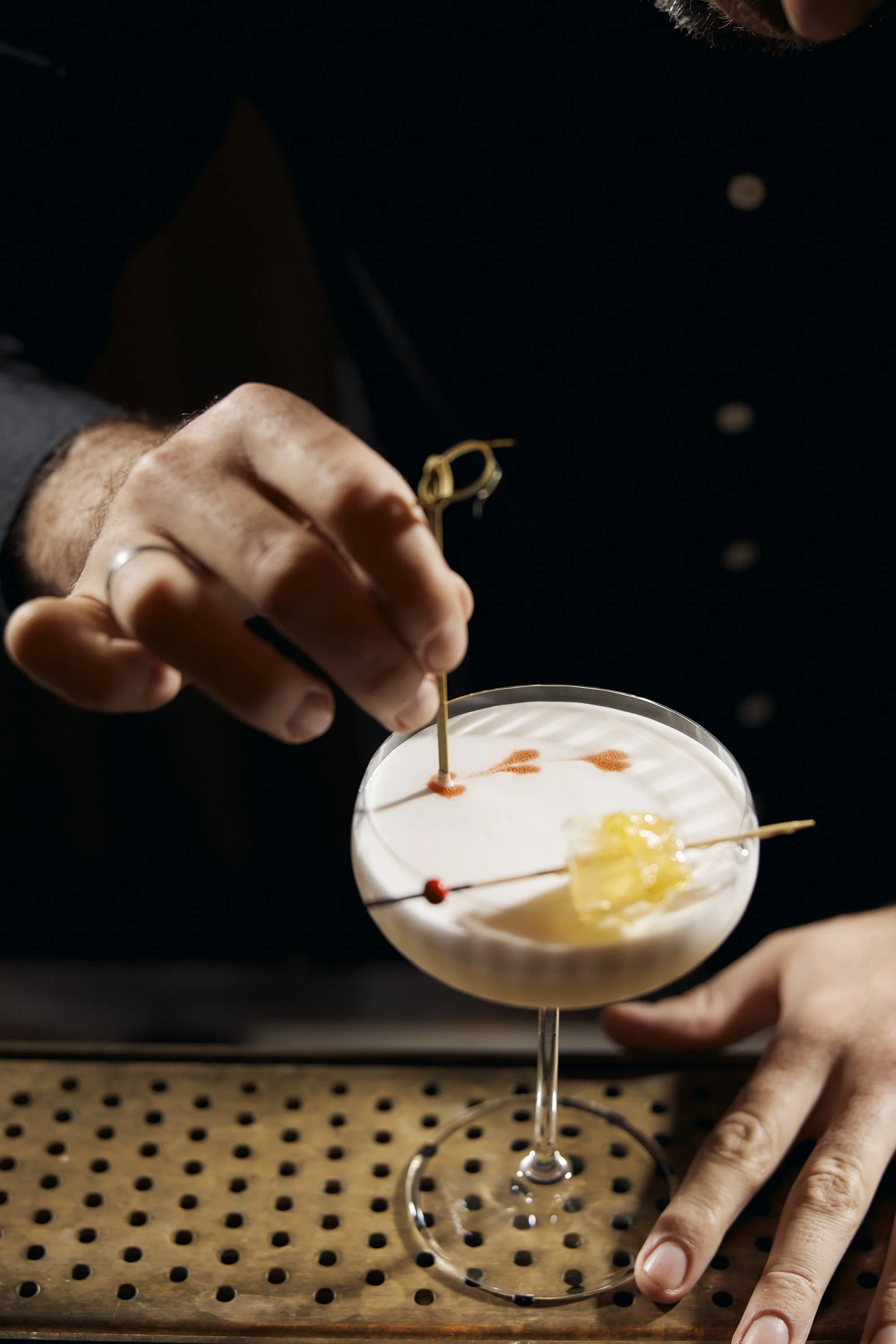 A bartender garnish a cocktail with a cherry and a citrus peel in a coupe glass. The bartender has a silver ring on their finger and is using tweezers.