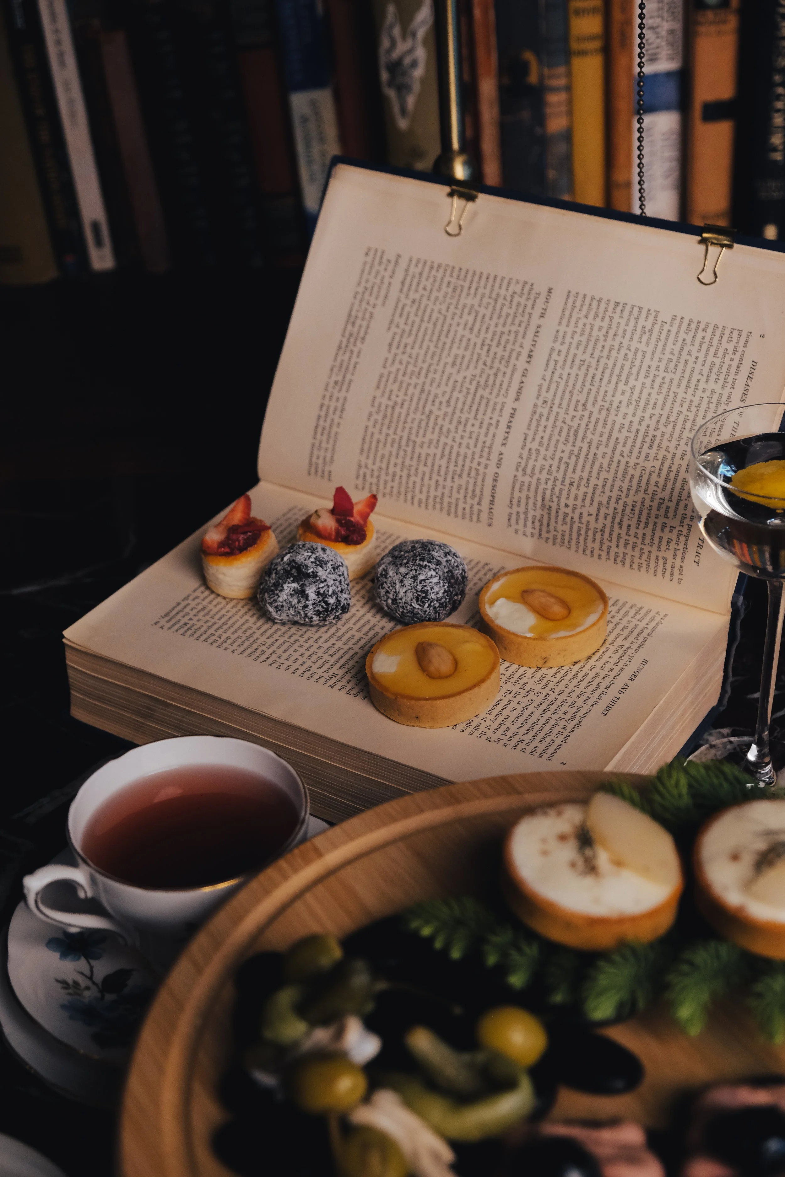 Open book with assorted desserts including fruit-topped tarts, chocolate truffles, and mini cakes, set on a table with a cup of tea, a glass of wine, and a wooden platter with cheese and vegetables.