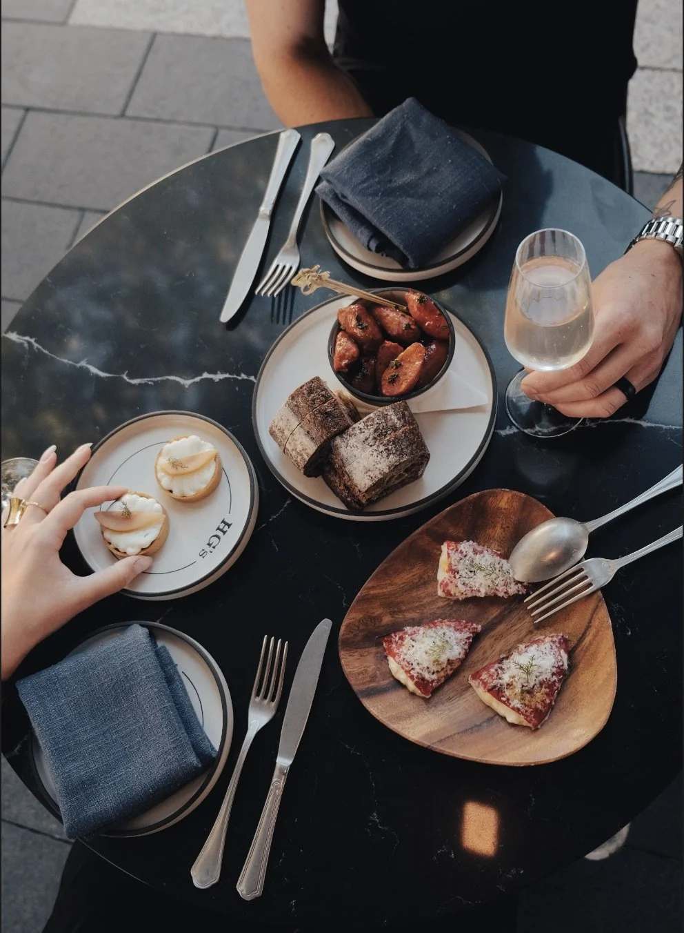 A table with assorted desserts and appetizers, including small tarts, chocolate cake rolls, a bowl of glazed sausages, and slices of pizza, with two people celebrating with drinks.