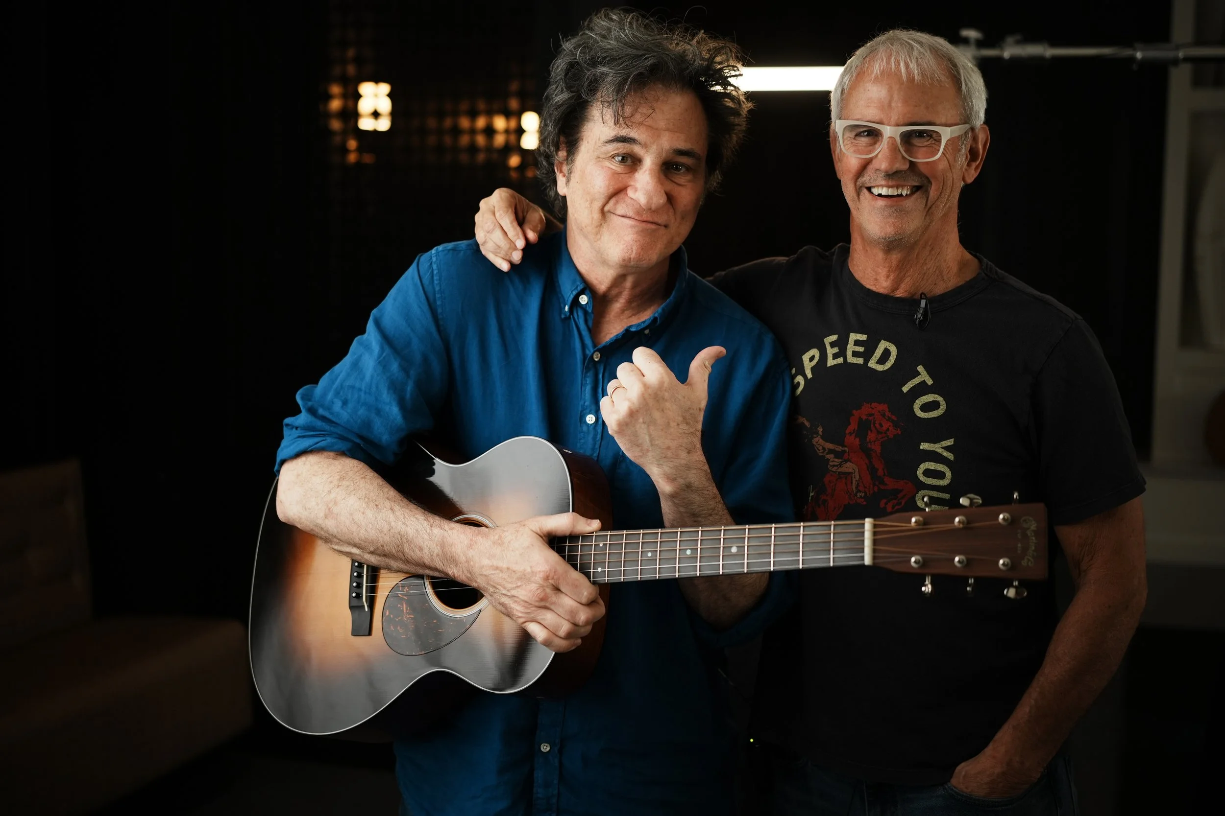 Two smiling men, one holding an acoustic guitar, standing close together with their arms around each other in a dark room with warm lighting.