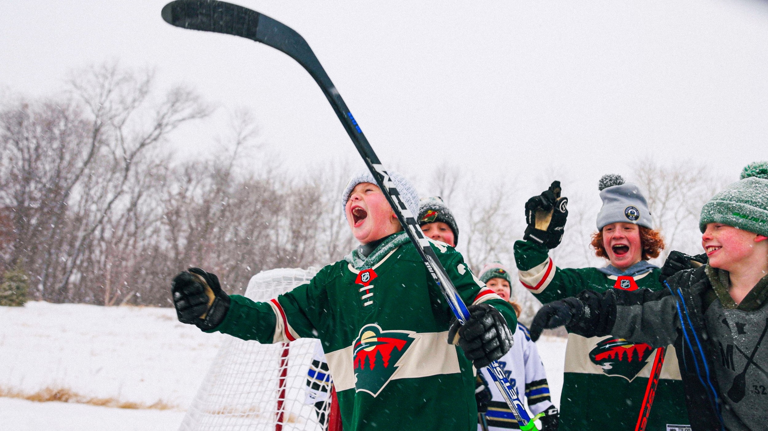 Group of children in hockey uniforms celebrating outdoors in snow, with one child holding a hockey stick aloft
