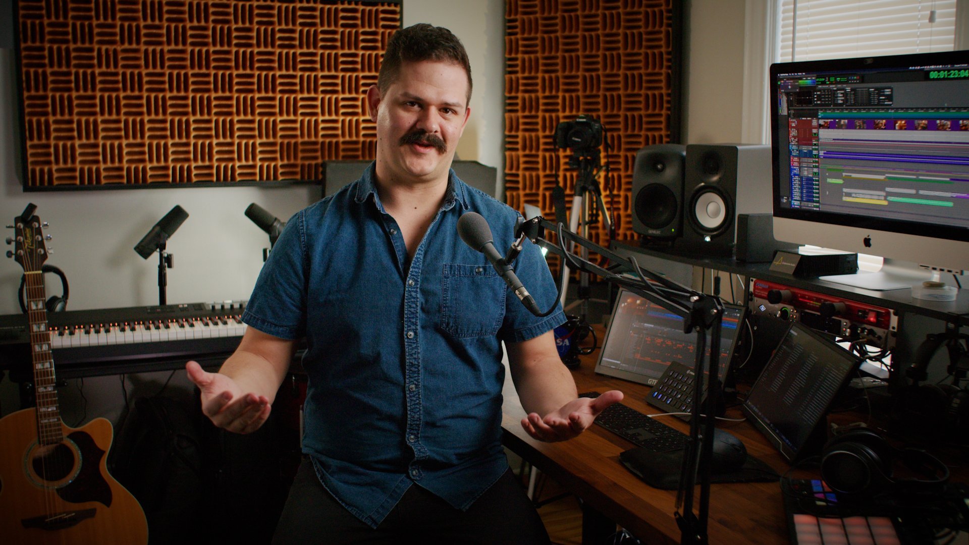 A man in a denim shirt talking in a recording studio, surrounded by music equipment including a guitar, MIDI keyboard, speakers, audio monitors, and multiple screens displaying digital audio workstations.